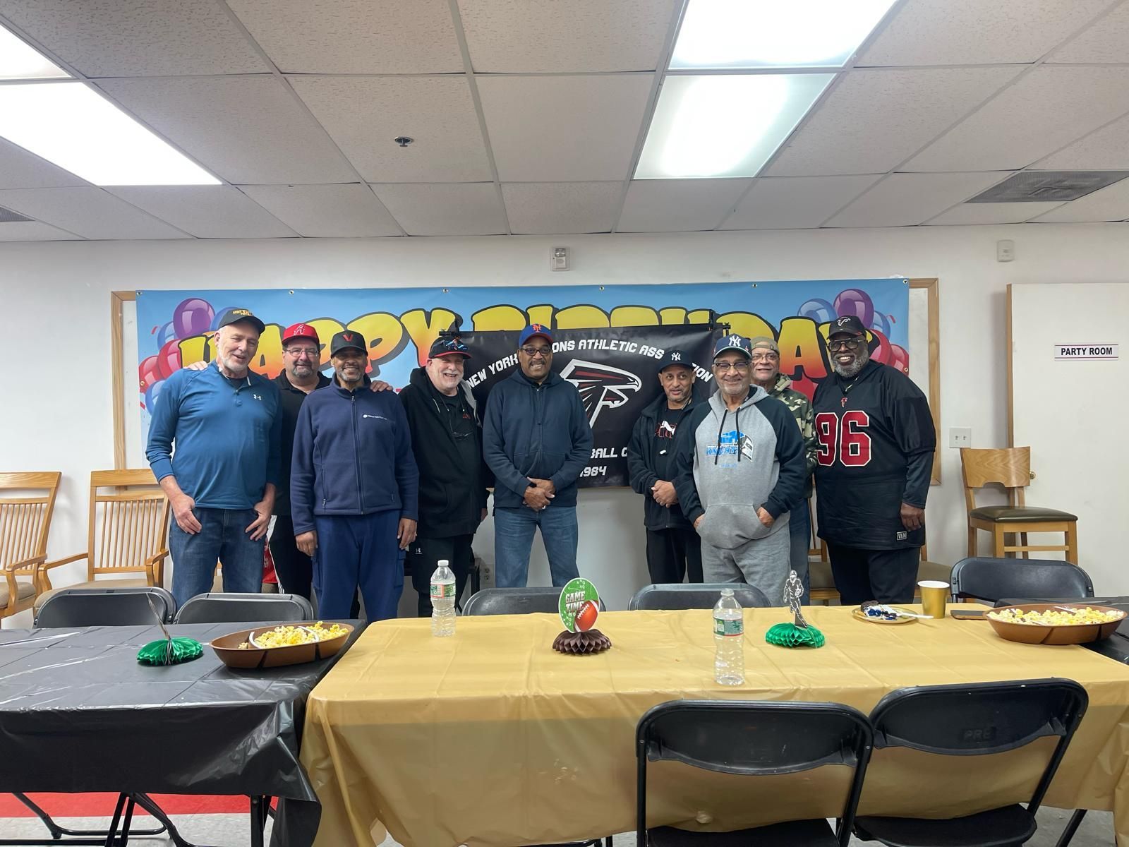 Group of people posing behind decorated tables in a community room with a colorful banner backdrop.