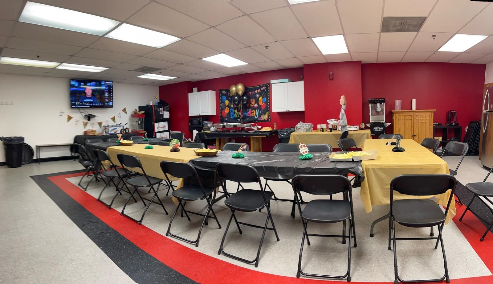 Community room with rows of folding chairs, long tables, and a wall-mounted TV on a red and white wall.