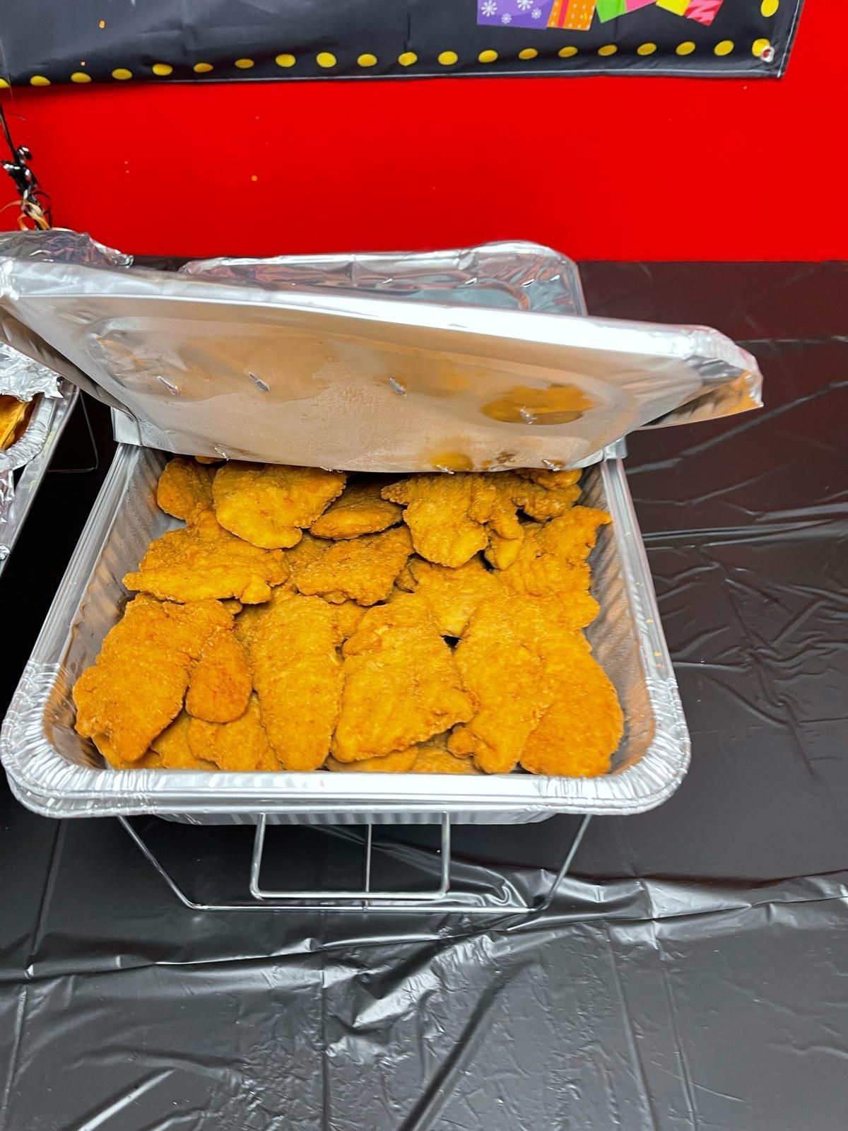 Tray of crispy fried chicken pieces in a foil pan, with a lid propped open at an outdoor setup