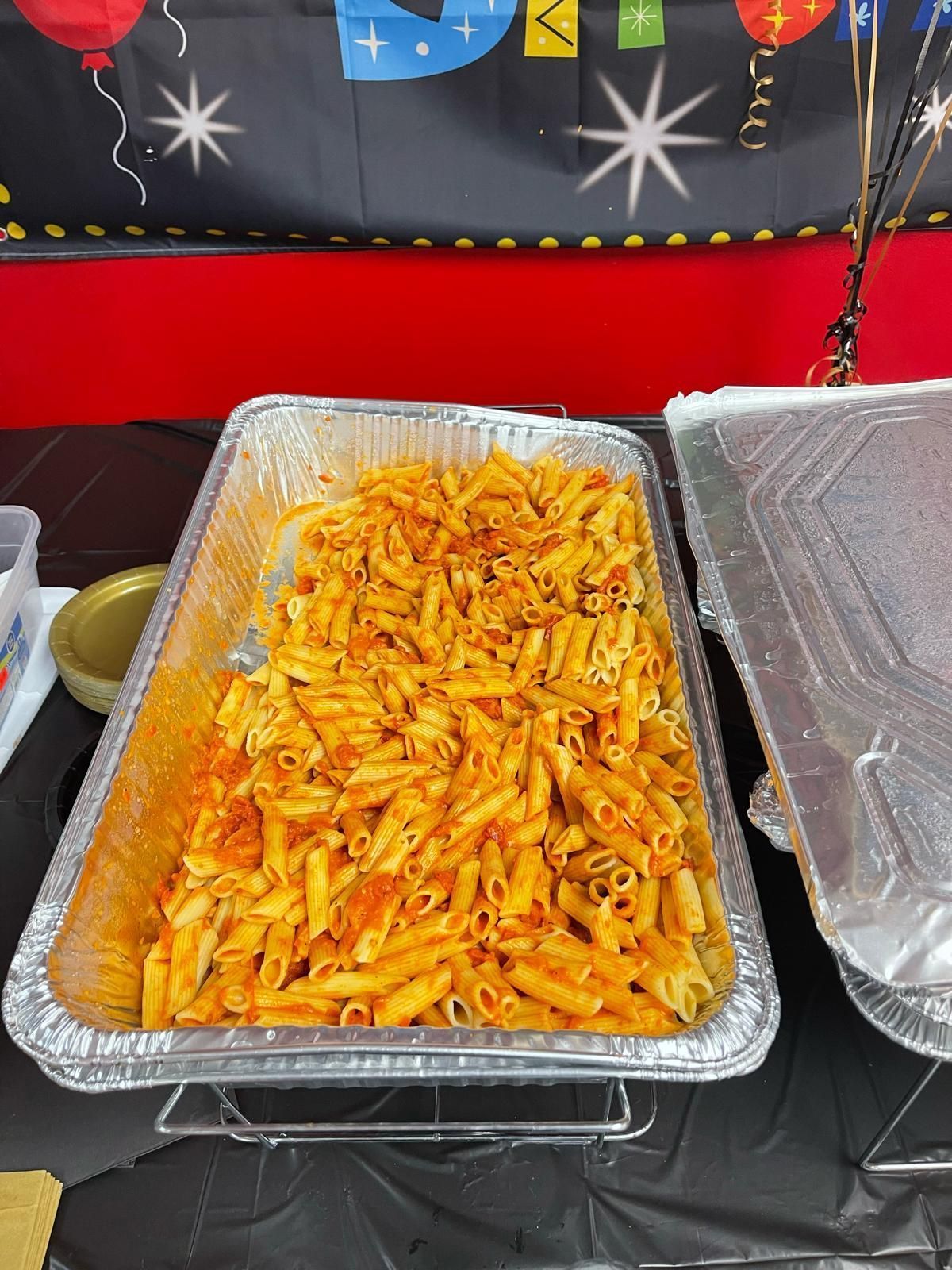 Tray of penne pasta in red sauce on a table, with covered side dishes nearby.