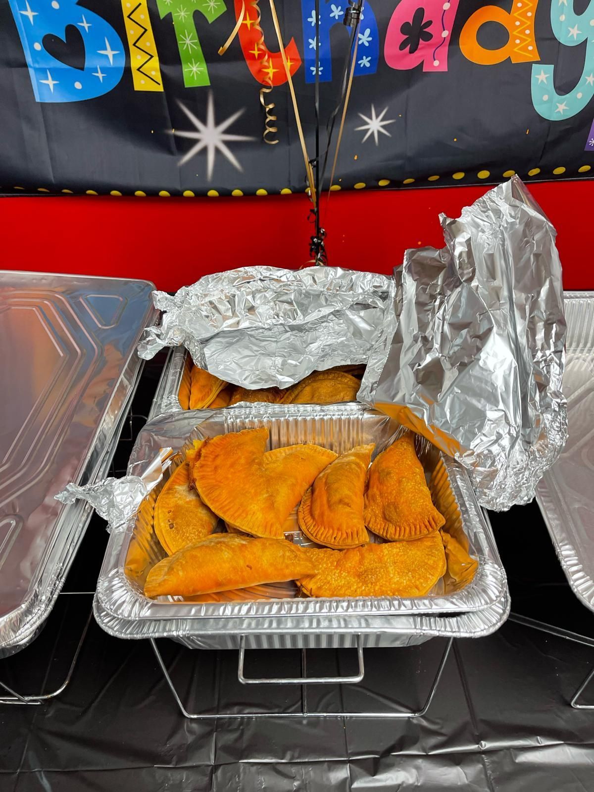 Foil takeout tray with fried food on a birthday-themed tablecloth, lid partially open.