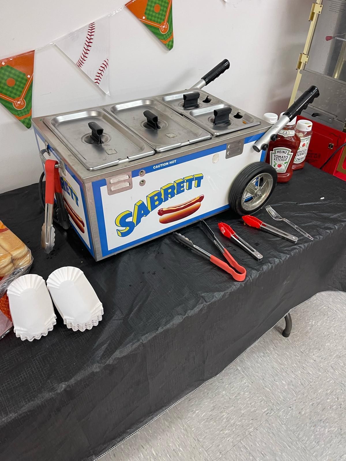 Stainless steel food cart with blue logo, utensils, and cups on a black tablecloth at an indoor event