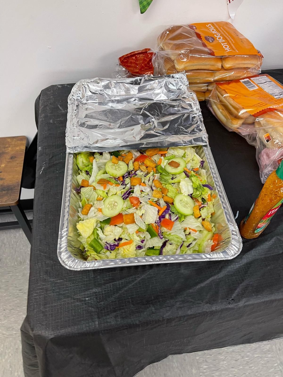 Foil tray of mixed salad on a black table, with wrapped bread or pastries stacked behind it