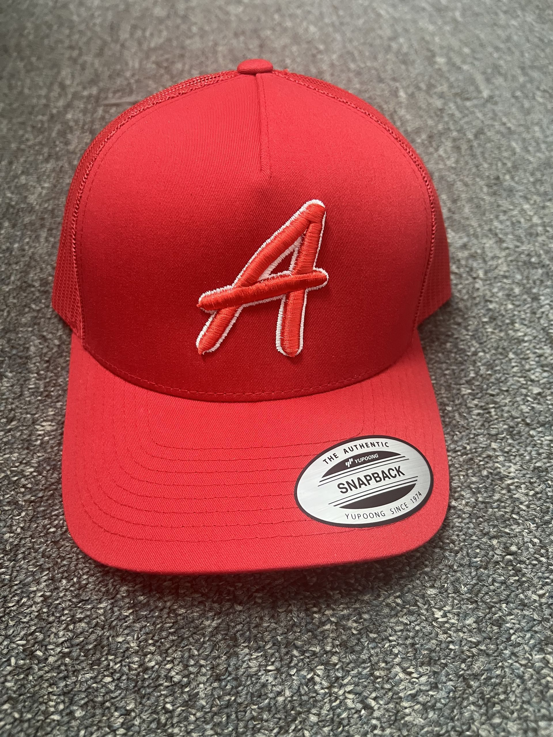 Red baseball cap with a white-outlined letter A on the front and a sticker on the brim.