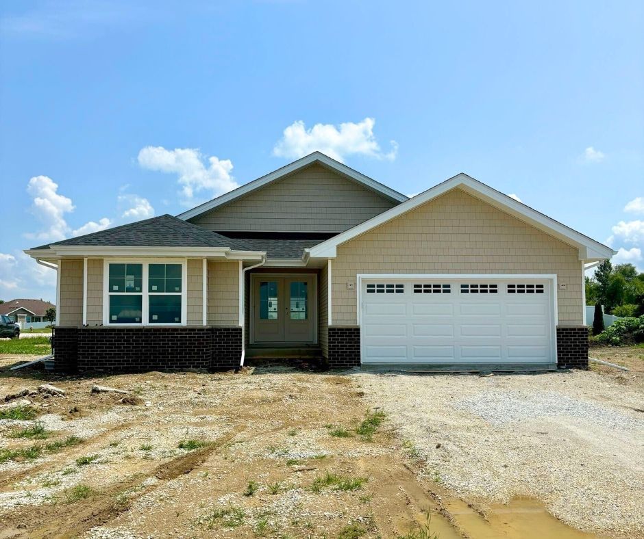 New tan and brown house with a white garage door and brick accents, set on a construction site under a blue sky.