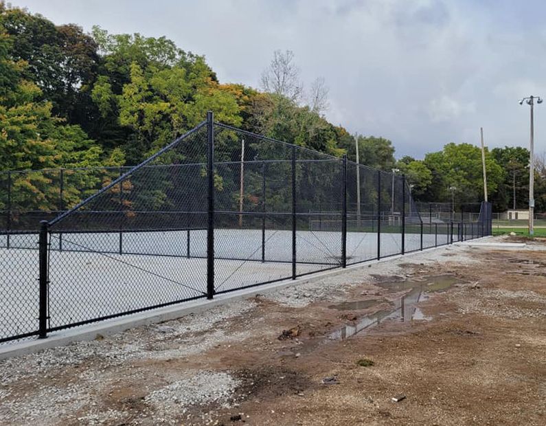 A chain link fence surrounds a tennis court in a park.