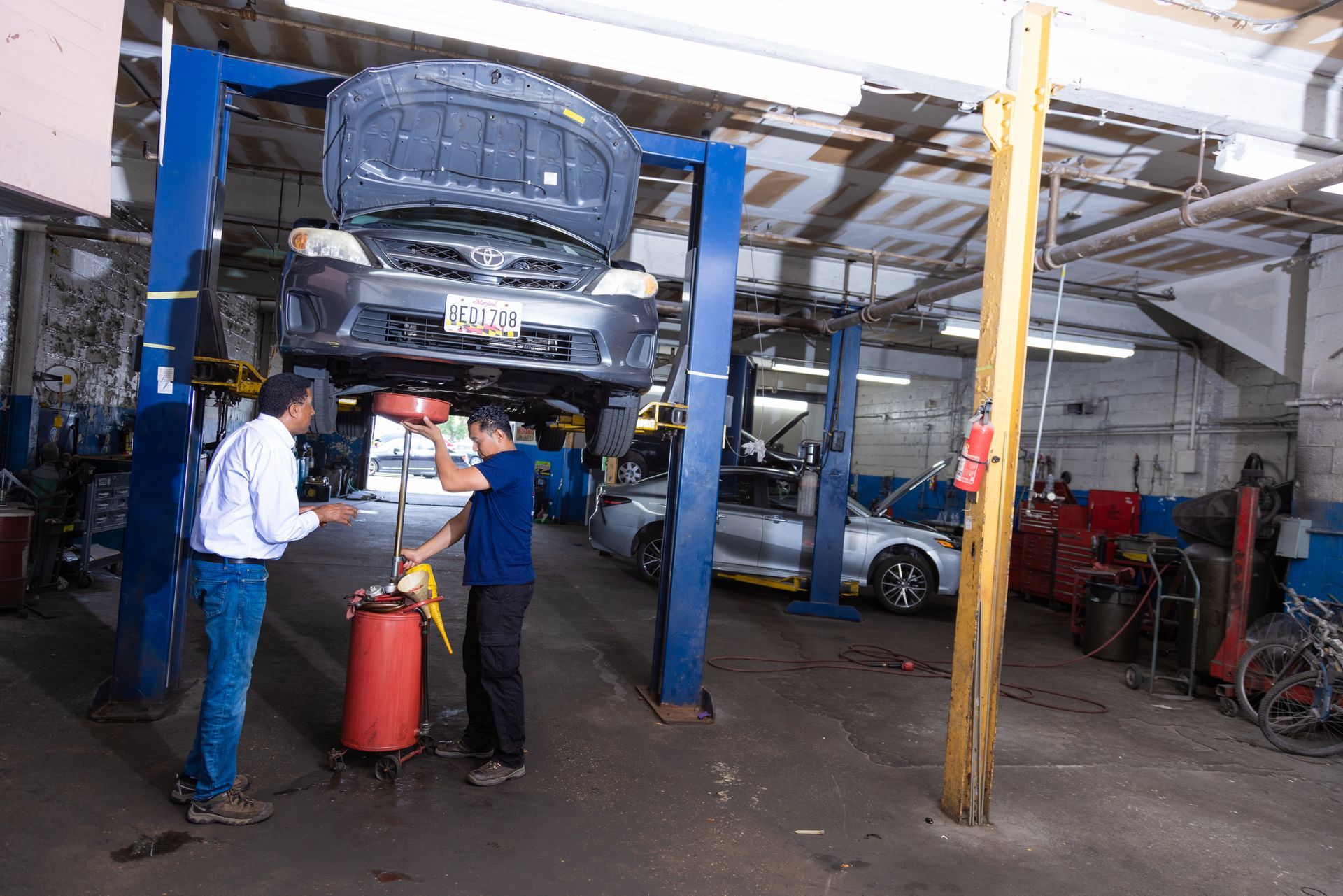 Two mechanics working in a garage; one holds a container while another is under a car raised on a lift.