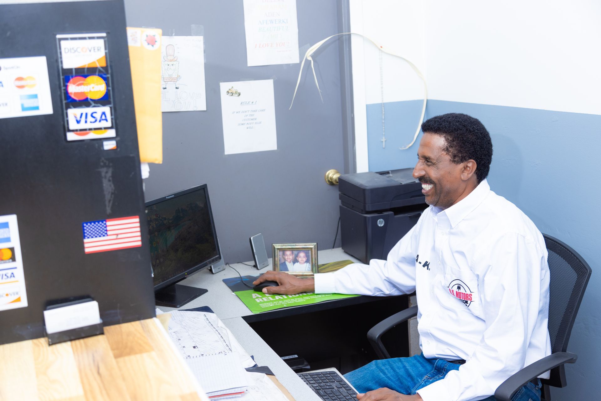 Man at desk smiling, using a computer. Credit card decals and American flag on nearby wall.