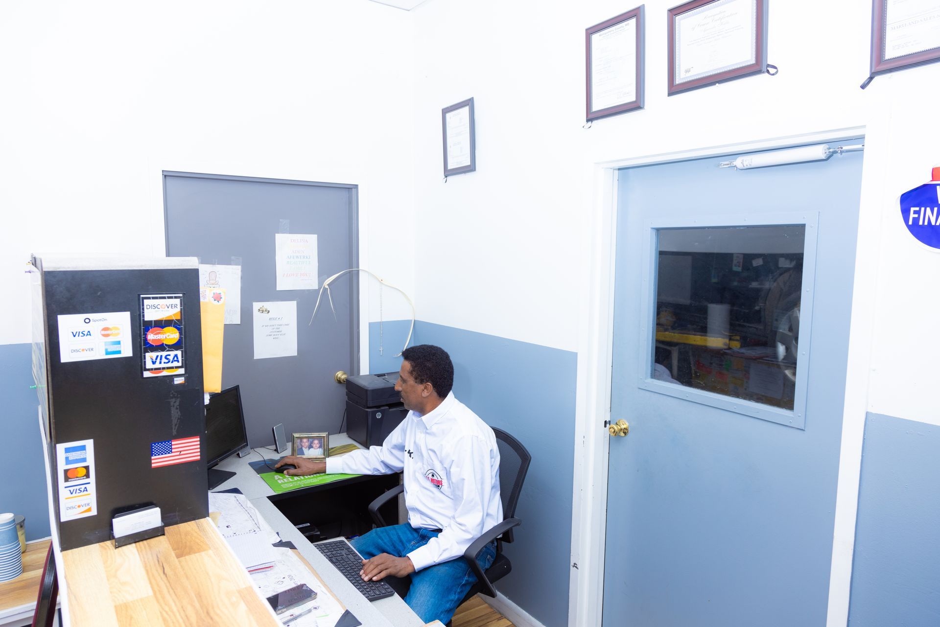 Man sits at a desk in a small office with computer, printer, and paperwork. Gray and blue walls, doors.