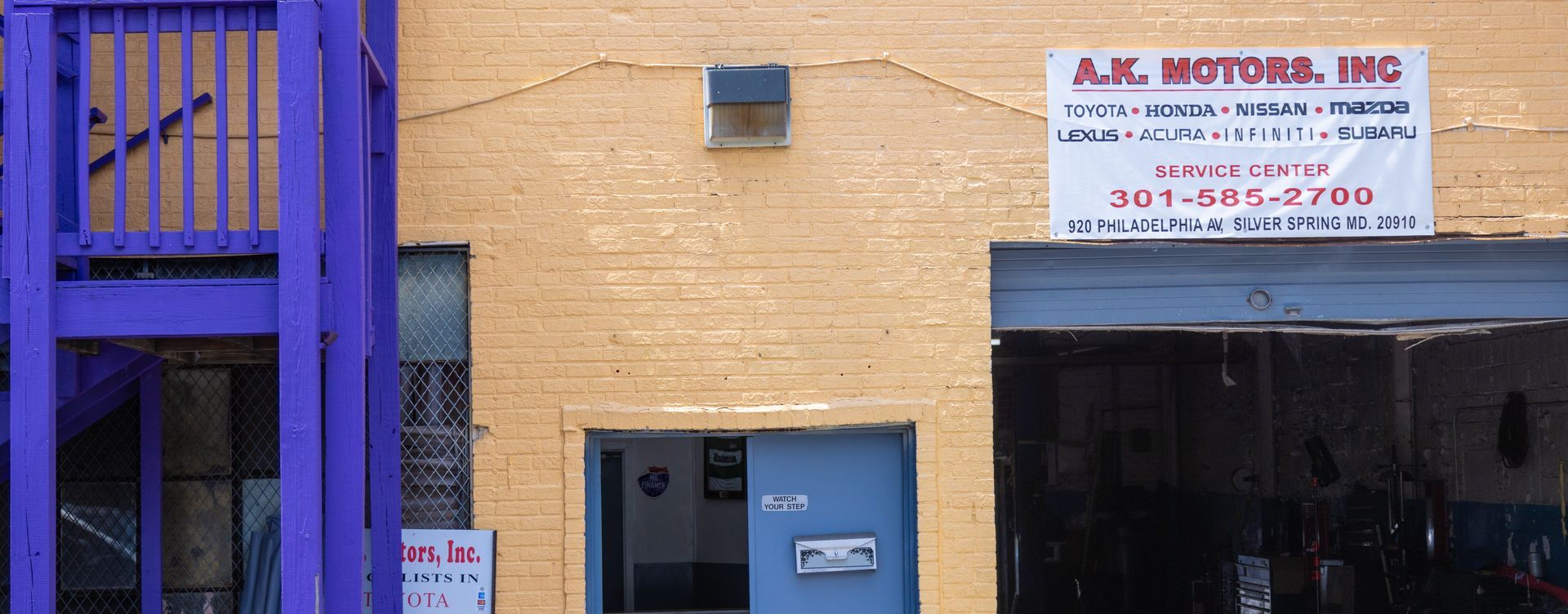 Exterior of J.K. Motors, Inc. building with blue door, purple scaffolding, and signage.