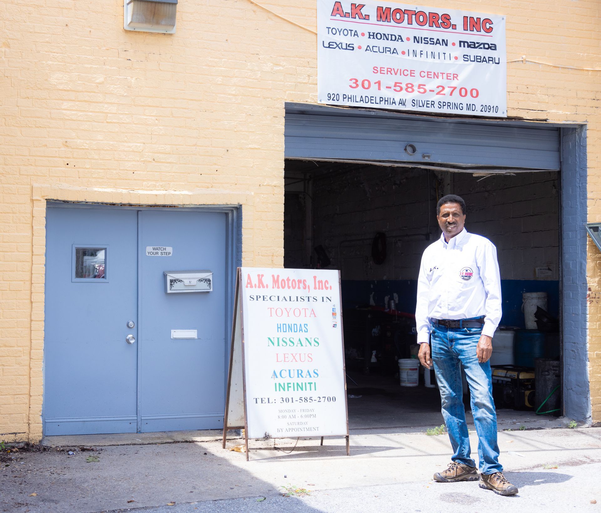 Man stands in front of A.K. Motors Inc., an auto repair shop with blue doors and an open garage.