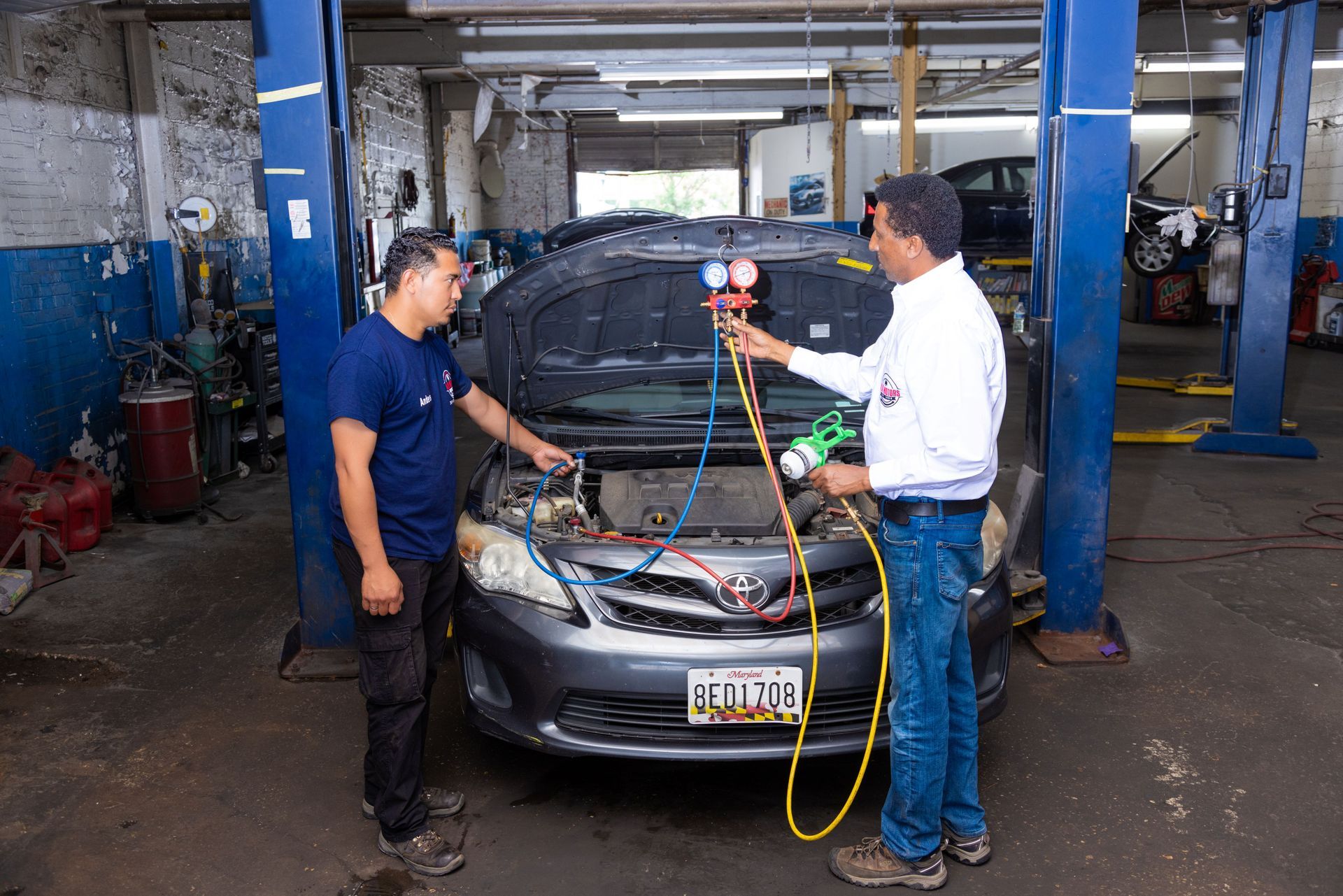 Two mechanics repairing a car's AC system in a garage. One holds gauges, other looks on.