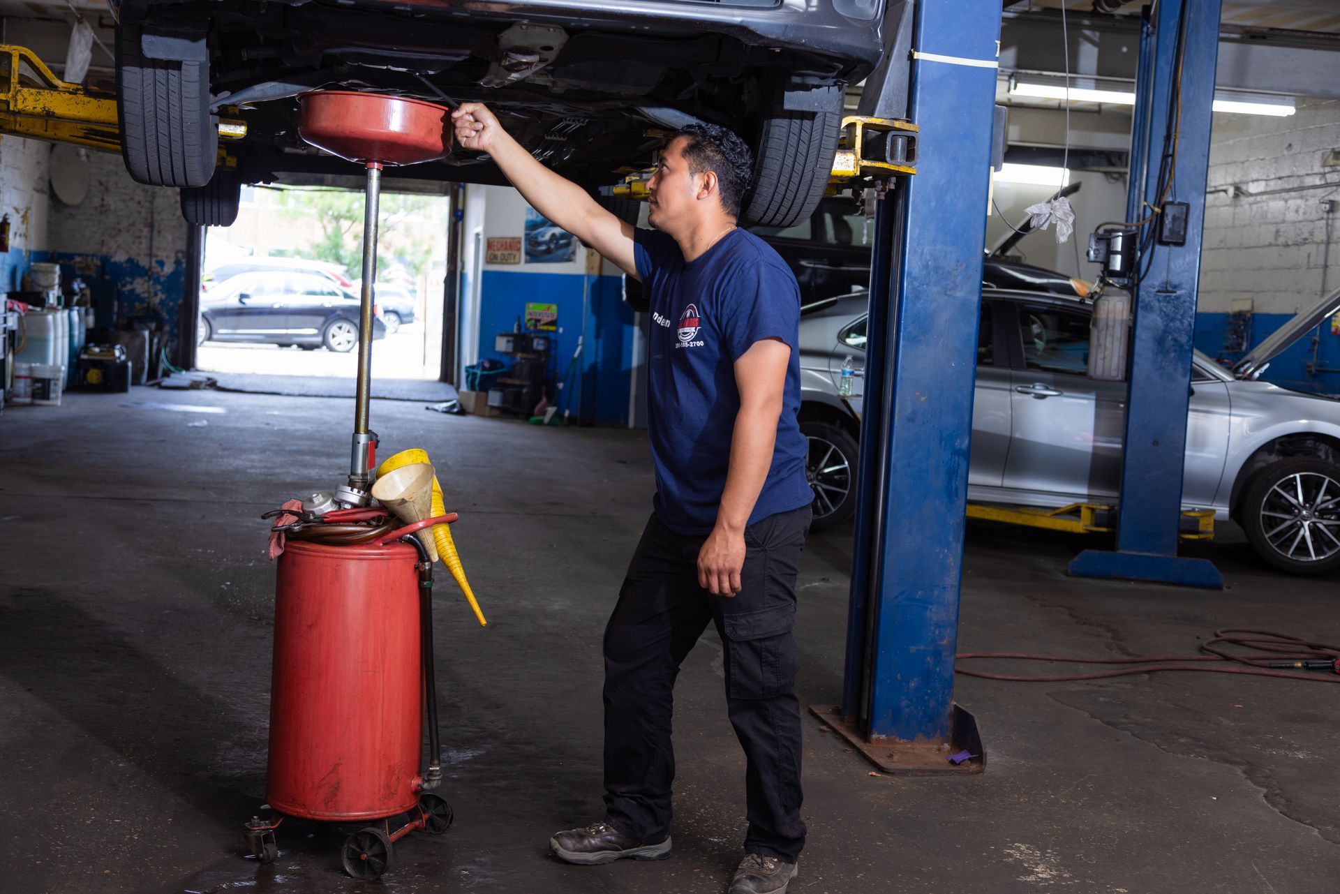 Mechanic draining oil from a car on a lift; wearing a blue shirt and black pants, in a garage.
