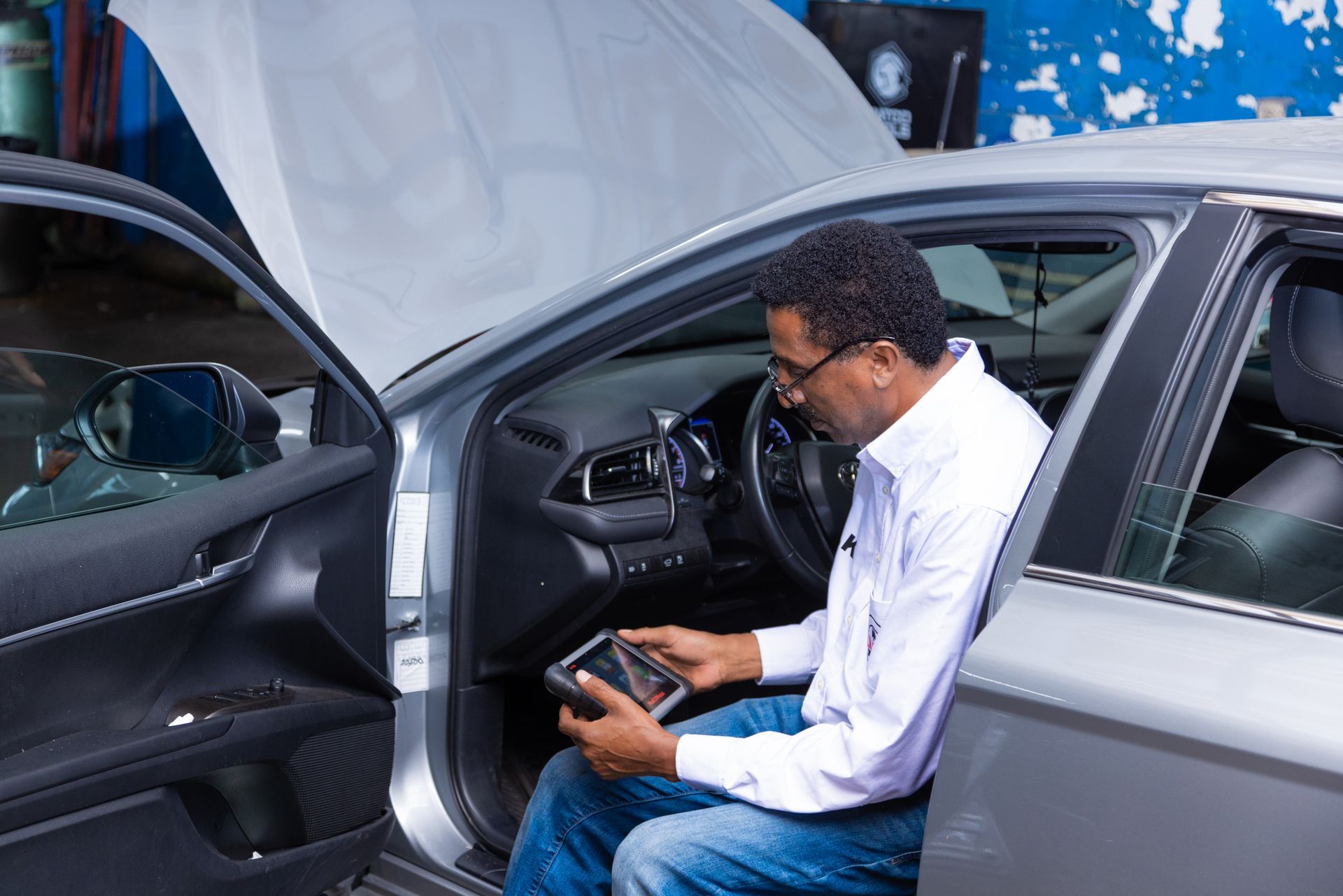 Man with glasses using a tablet inside a car with the hood open in a garage.