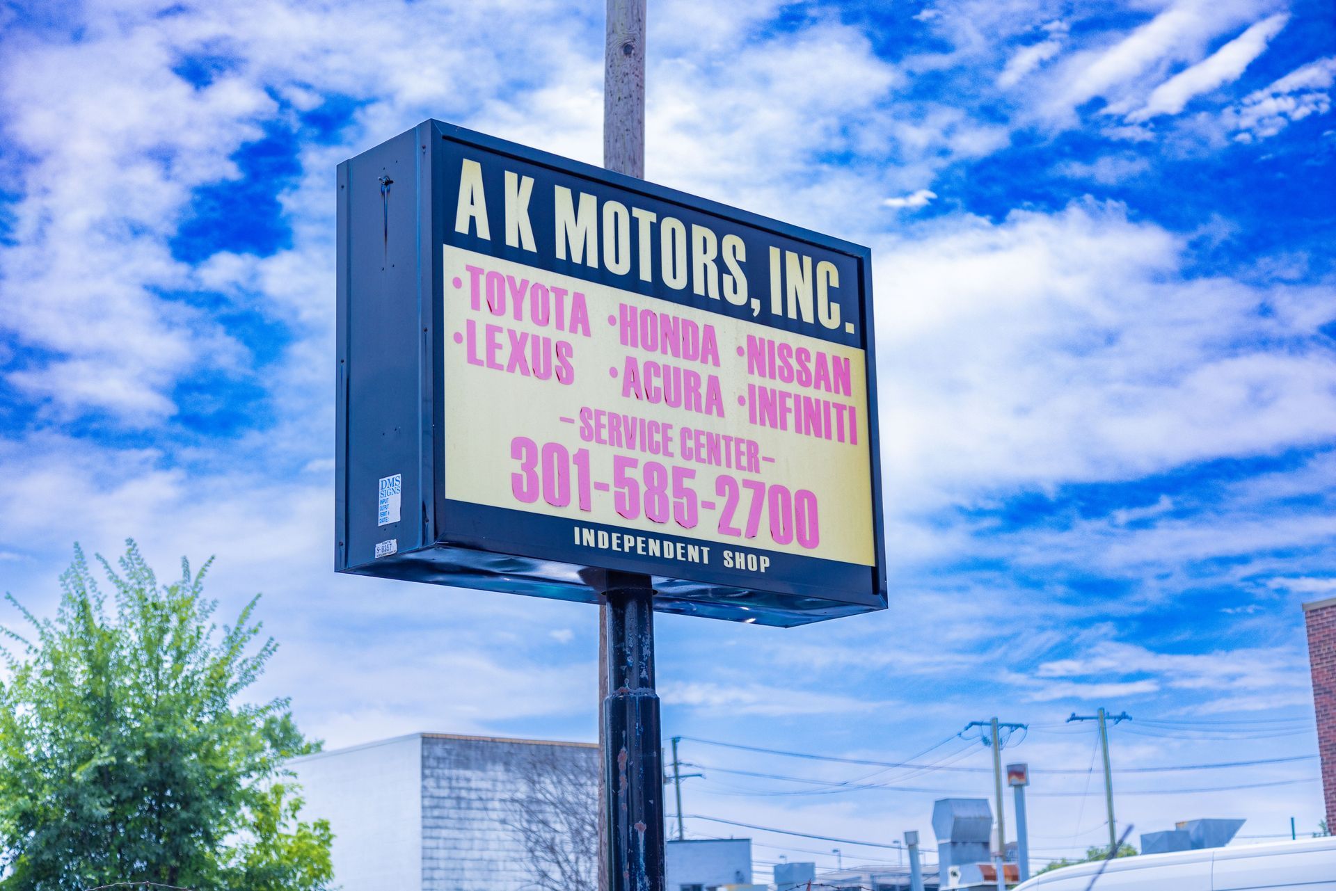 Sign for AK Motors, Inc. listing Toyota, Honda, Nissan, Lexus, Acura, Infiniti service; phone number visible against cloudy sky.
