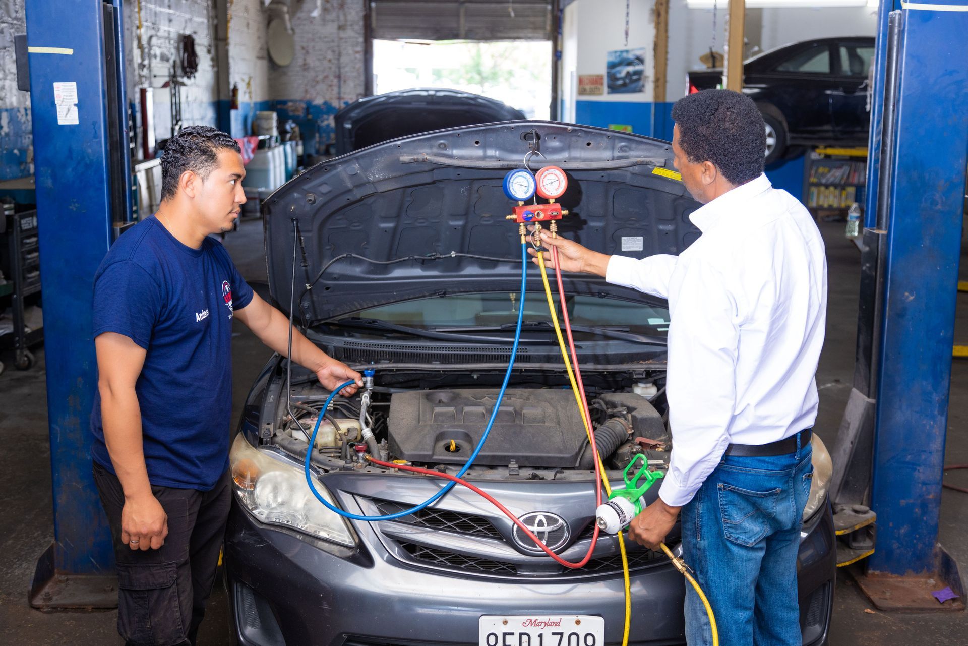 Two mechanics servicing a car's engine using gauges in a garage, one holding the device, the other assisting.
