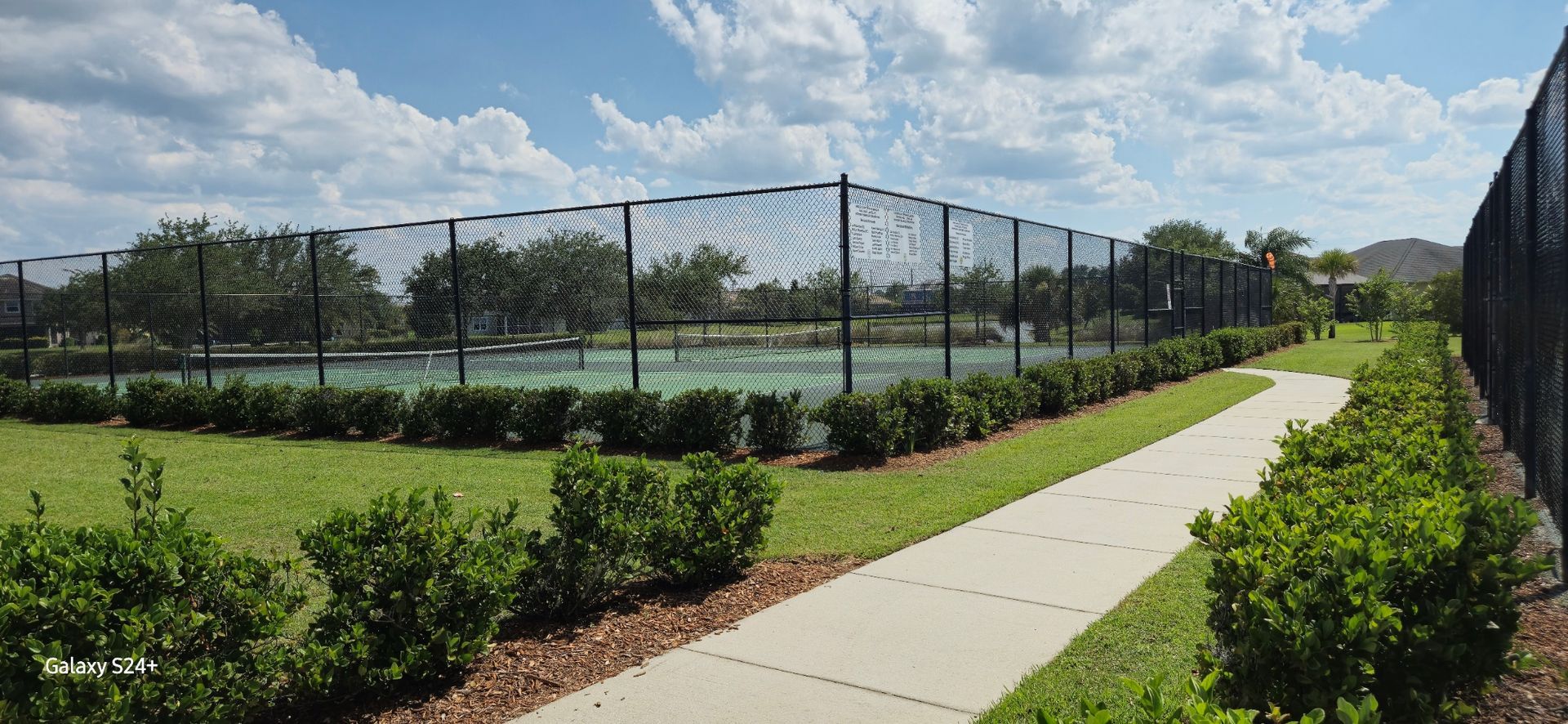 A tennis court is surrounded by a fence and a sidewalk.