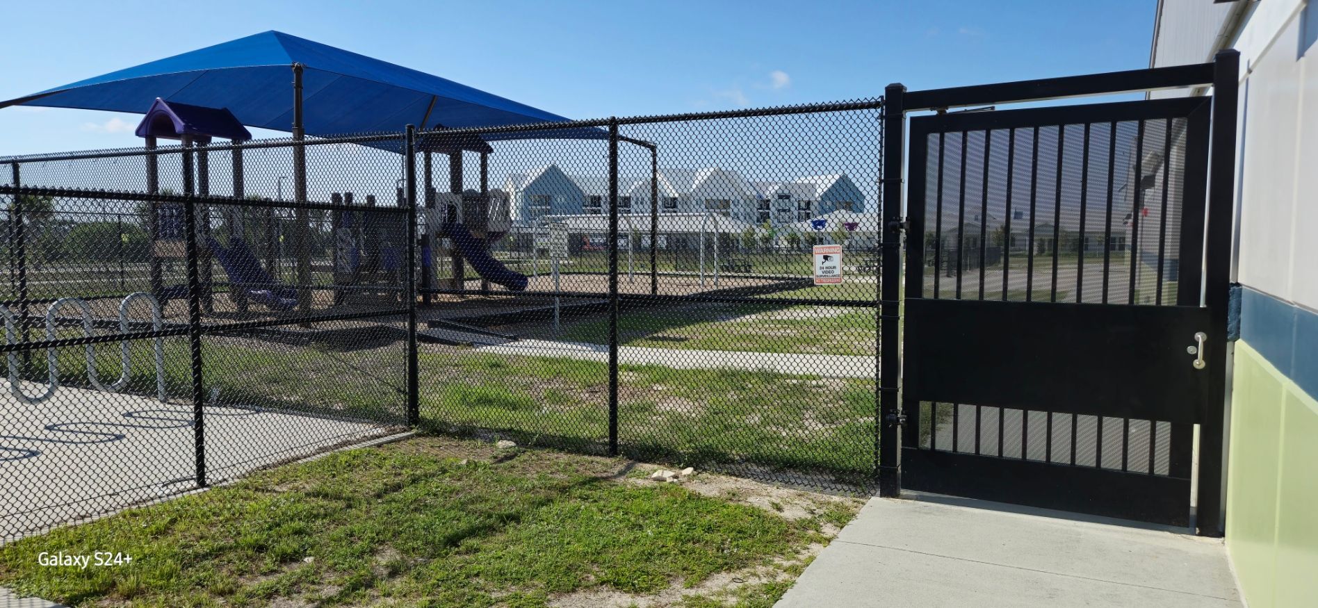 A chain link fence surrounds a playground with a blue umbrella.