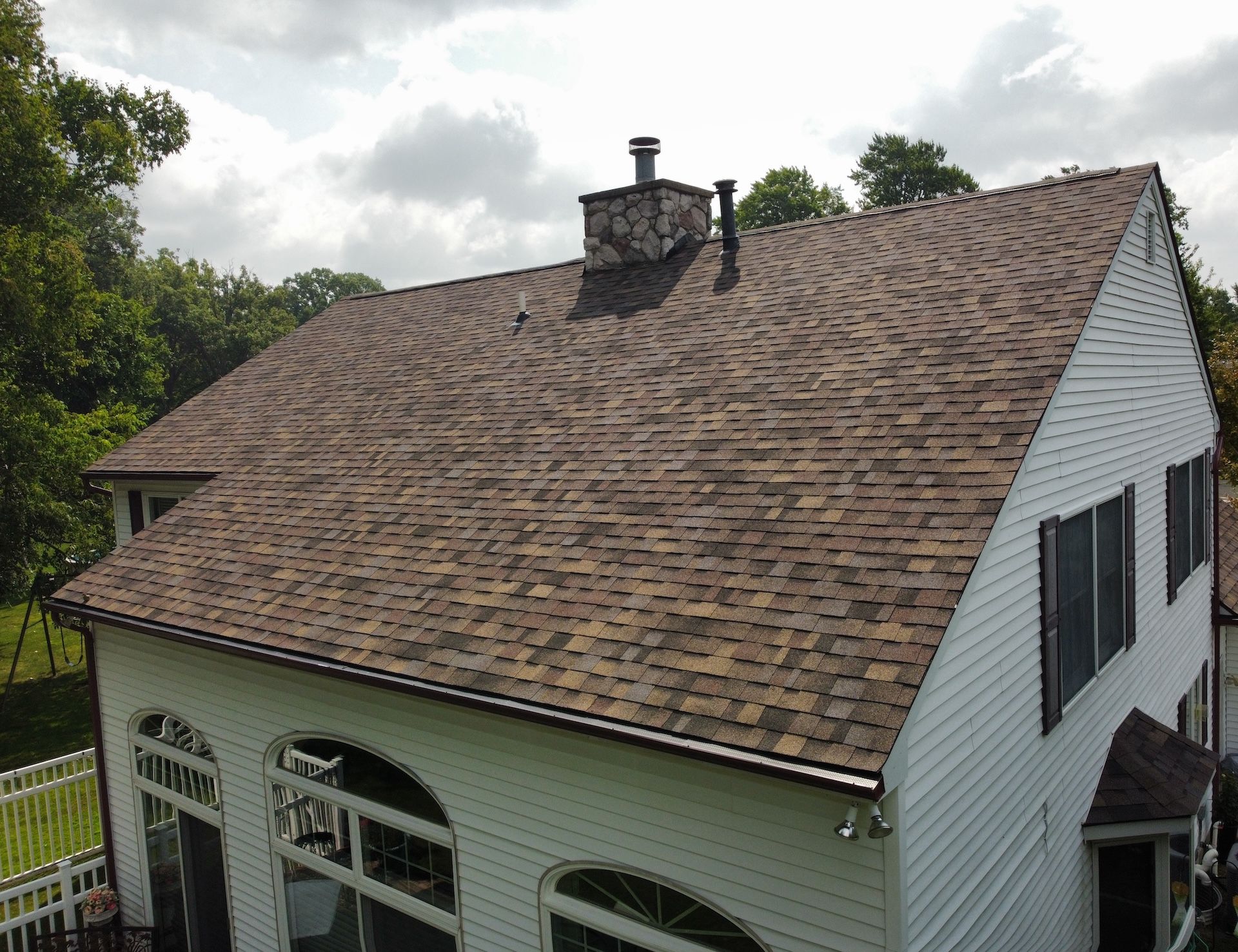 House roof with brown shingles and a central chimney, viewed from above under cloudy skies