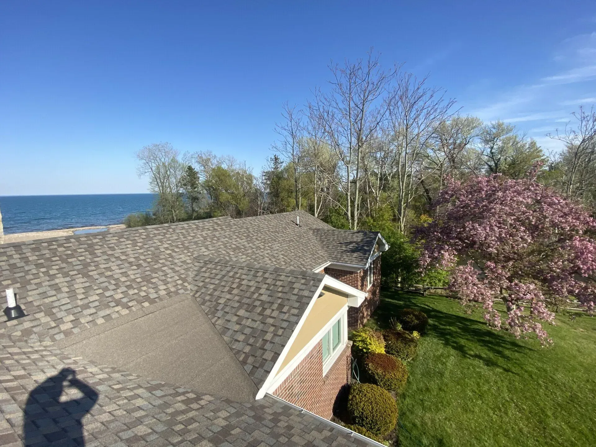 Lakefront house roof beside blue water, with trees and pink blossoms under a clear sky.