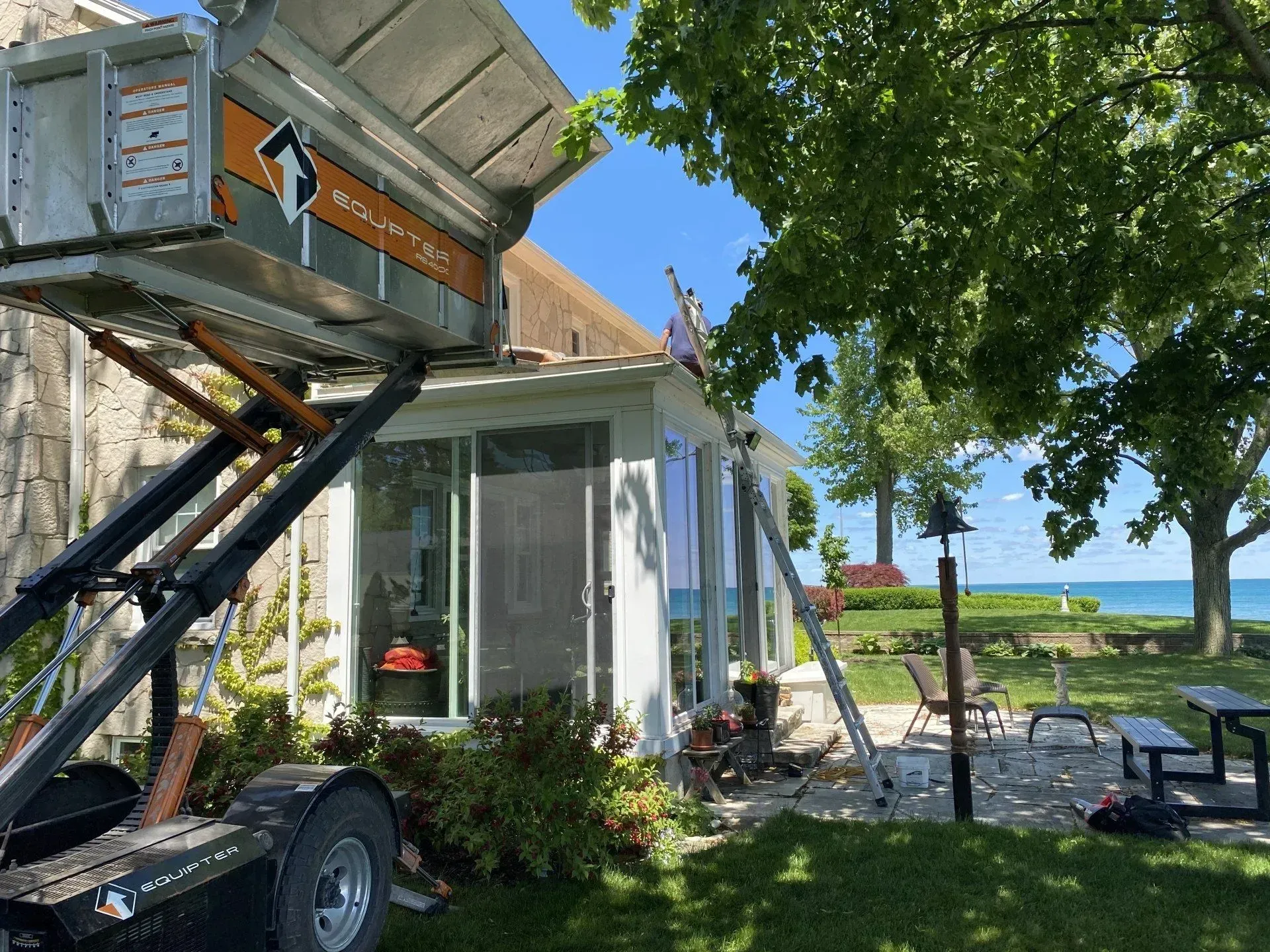Outdoor patio beside a white house with a scissor lift, tree shade, and a view of the water beyond.
