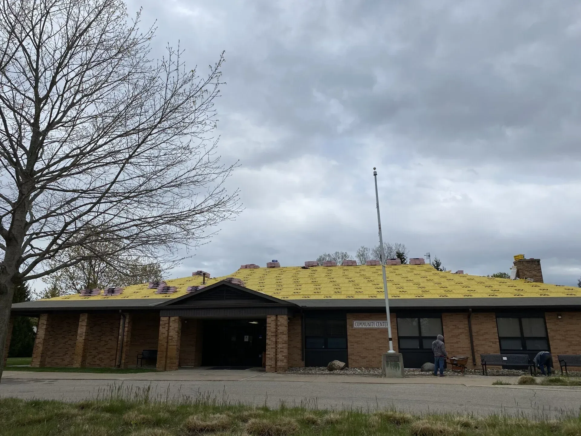 Low brick building with a yellow roof under gray clouds, tree at left, and a person near the entrance.