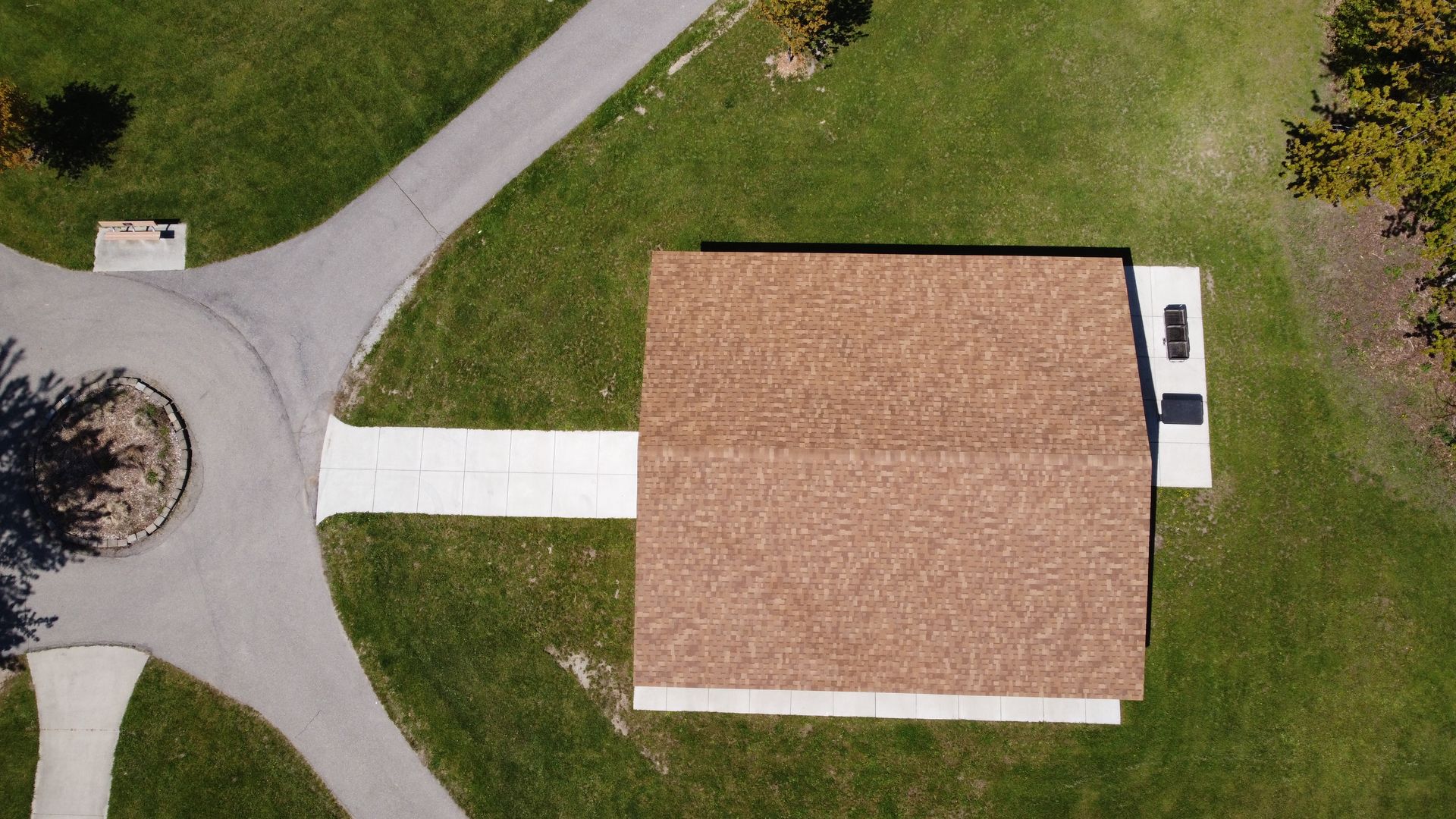 Aerial view of a tan-roofed house with a white driveway beside a curved road and green lawn.