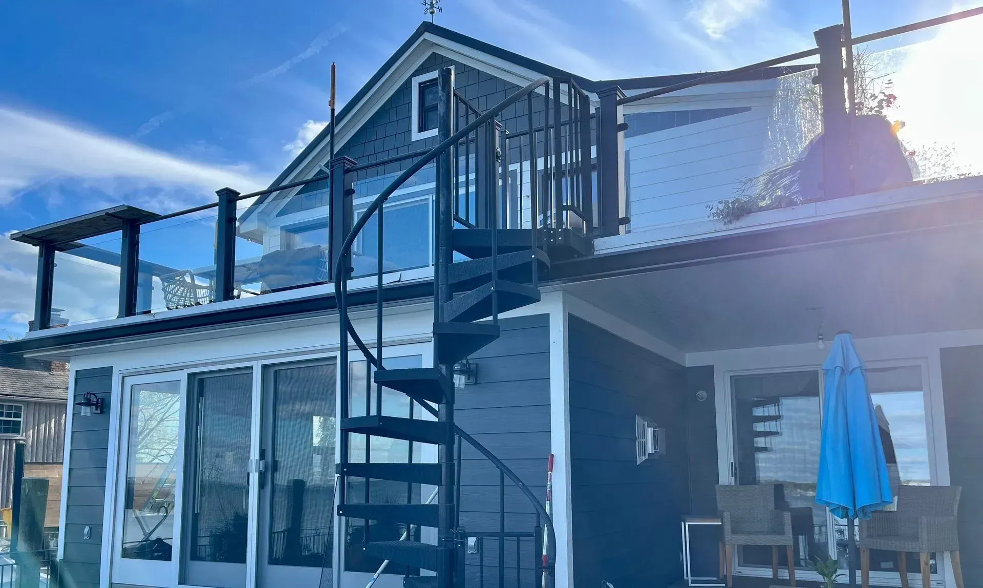 Two-story house with a black metal exterior staircase, glass railing, and a blue patio umbrella in bright sunlight