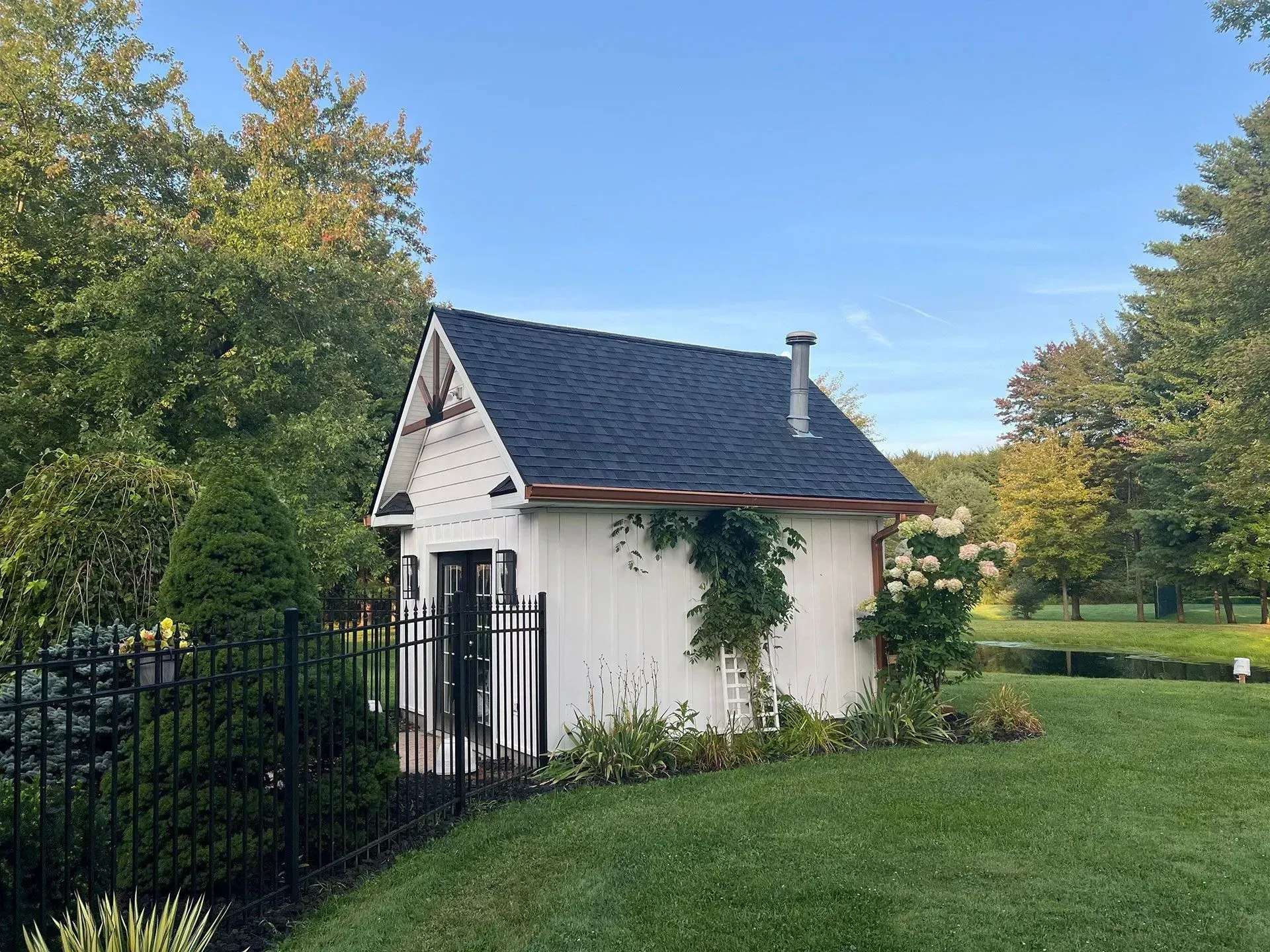 Small white shed with dark roof and black gate, set in a grassy yard with trees and blue sky.