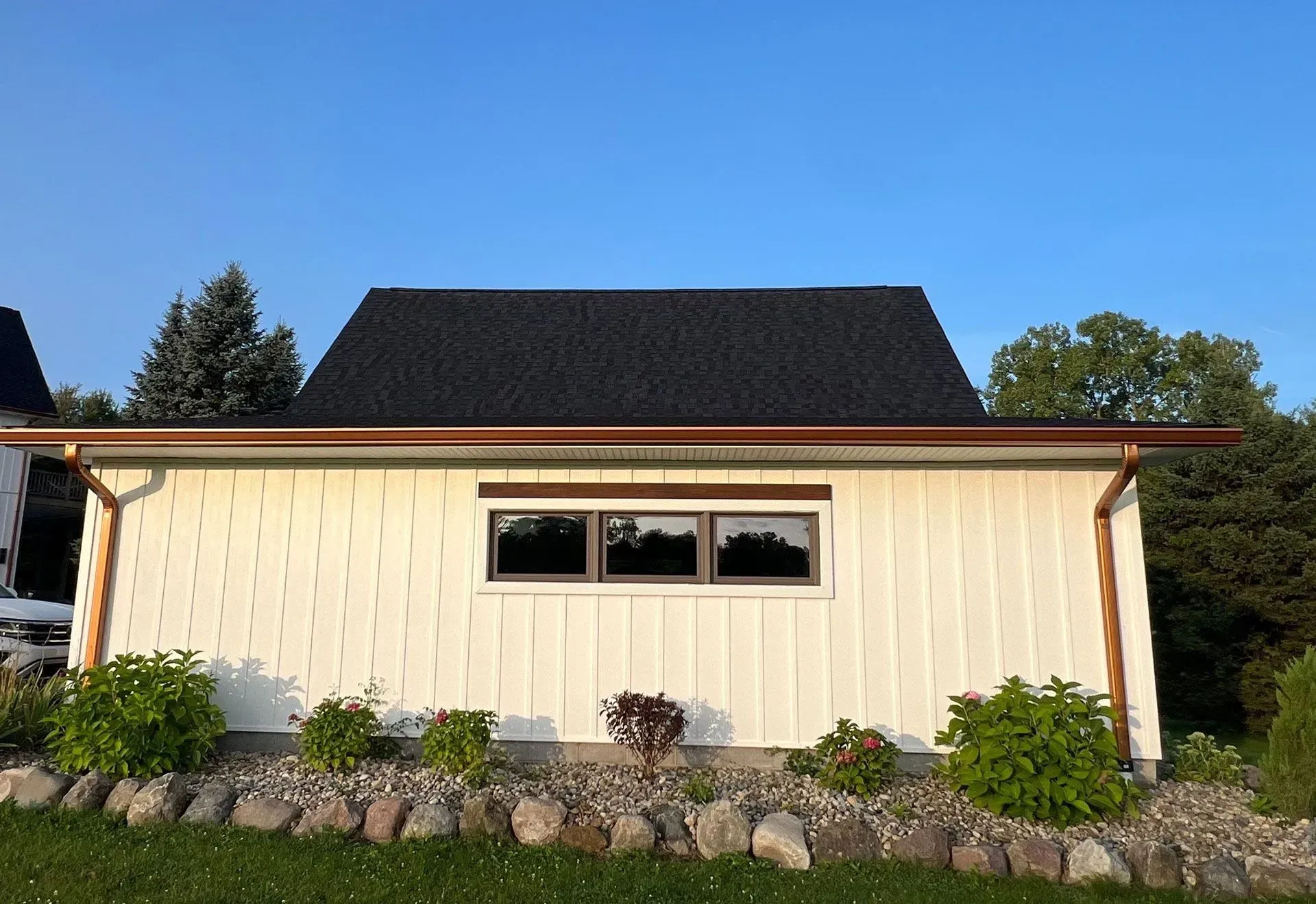 Cream-colored house exterior with dark roof, front window, and shrubs along a stone border.