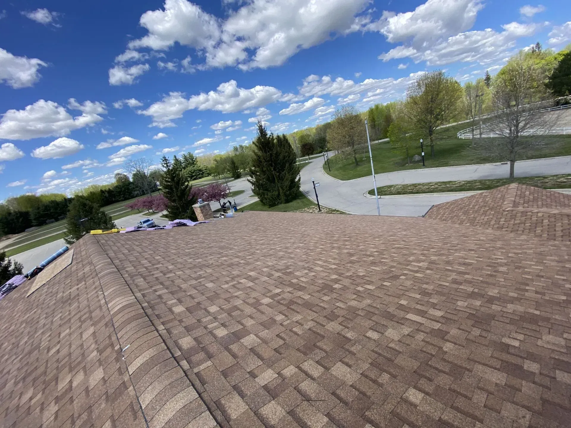 View from a shingled rooftop over a road, trees, and parked cars under a bright blue sky with clouds