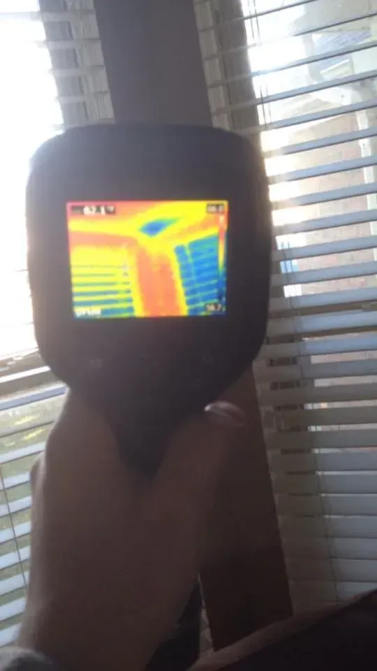 Handheld thermal camera displaying a colorful heat map in front of window blinds