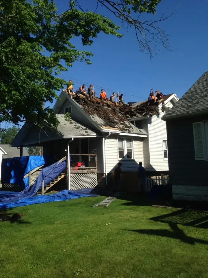 Roofing crew repairing storm-damaged house roof with exposed rafters and debris under a clear blue sky