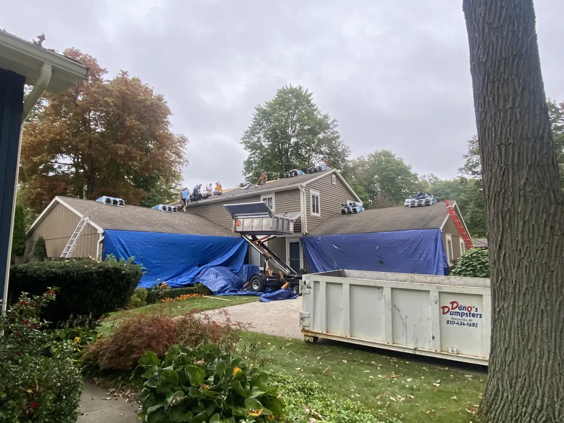 Collapsed house under repair with blue tarps and workers on the roof, debris and a dumpster in front
