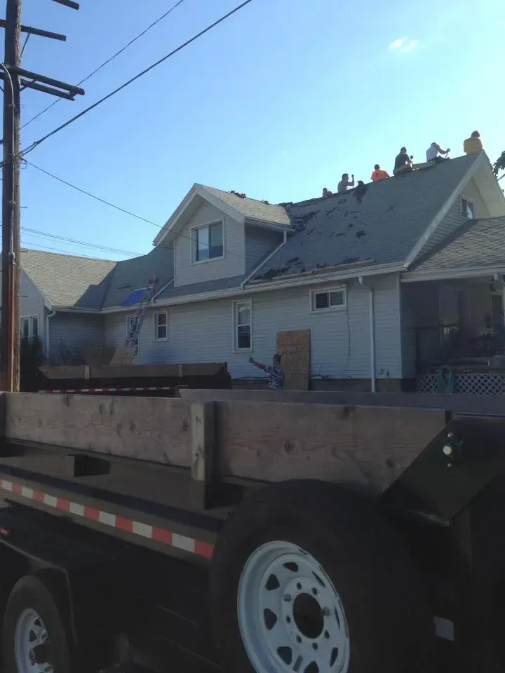 Flatbed truck carrying roofing materials beside a house with workers on the roof.