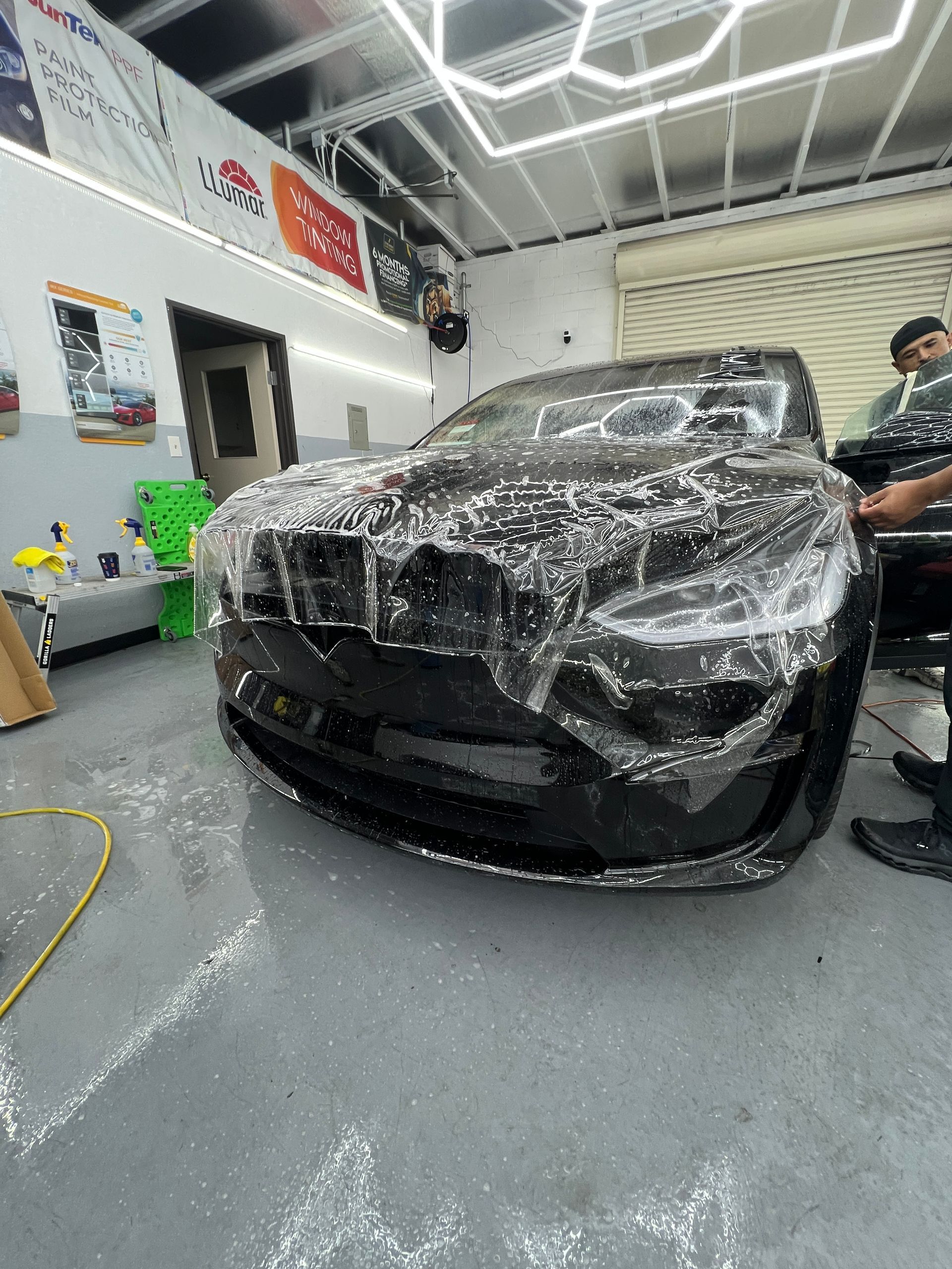 Black car wrapped in protective plastic in an auto workshop, with a person standing nearby