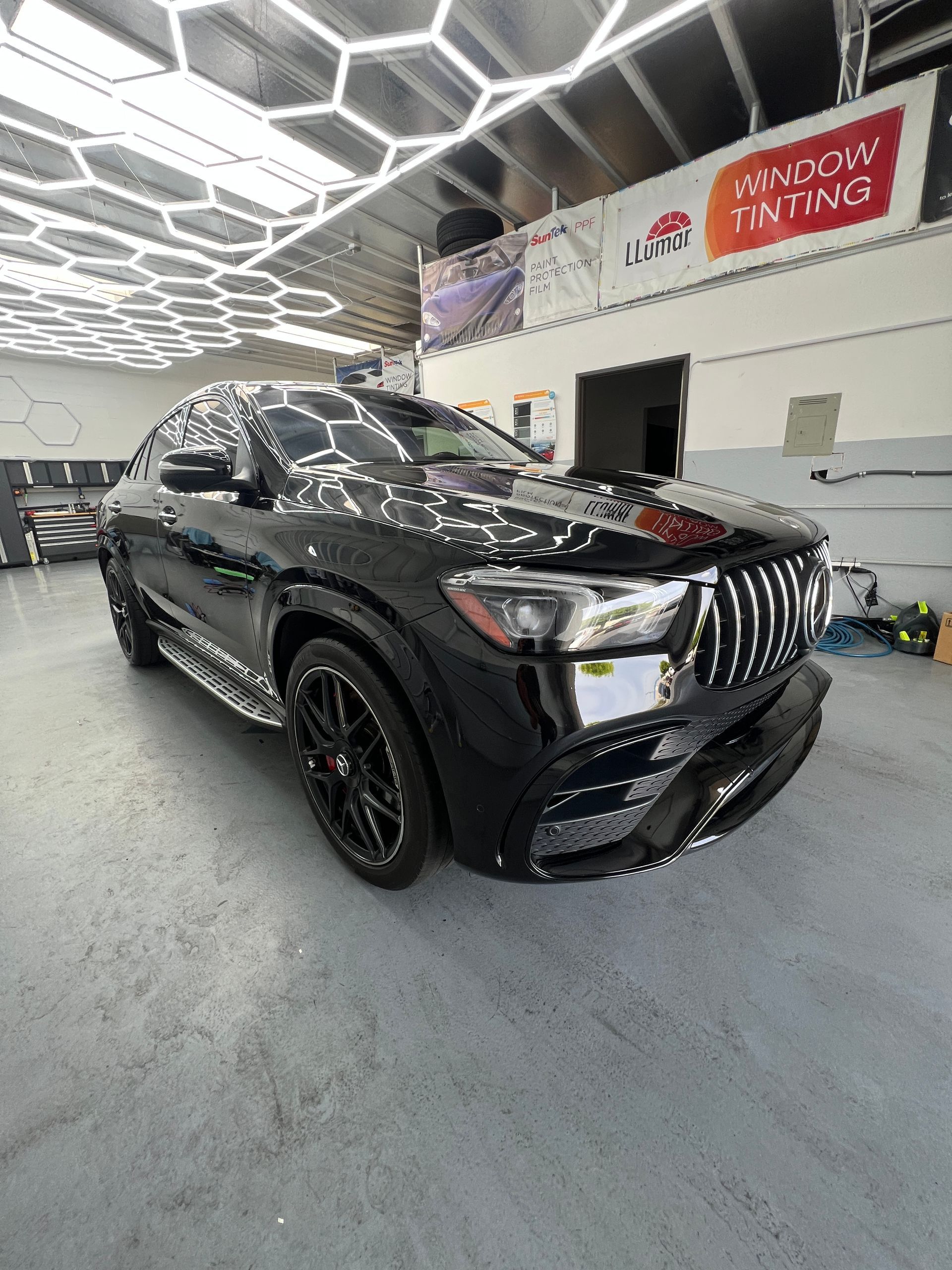 Black SUV in an auto detailing shop with bright hexagonal ceiling lights and a window tinting sign.