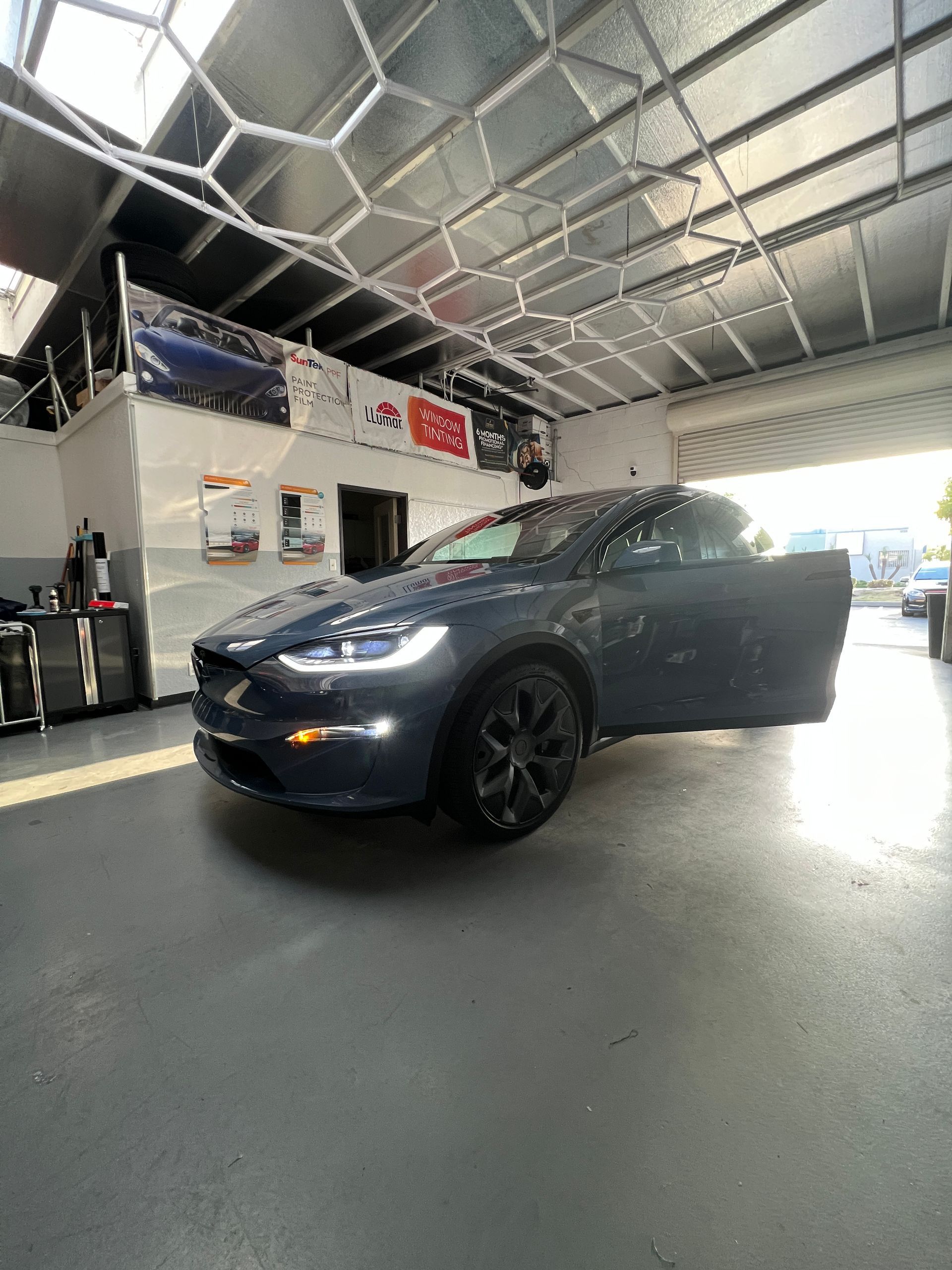 Blue Tesla sedan inside a garage with its headlights on and front passenger door open.