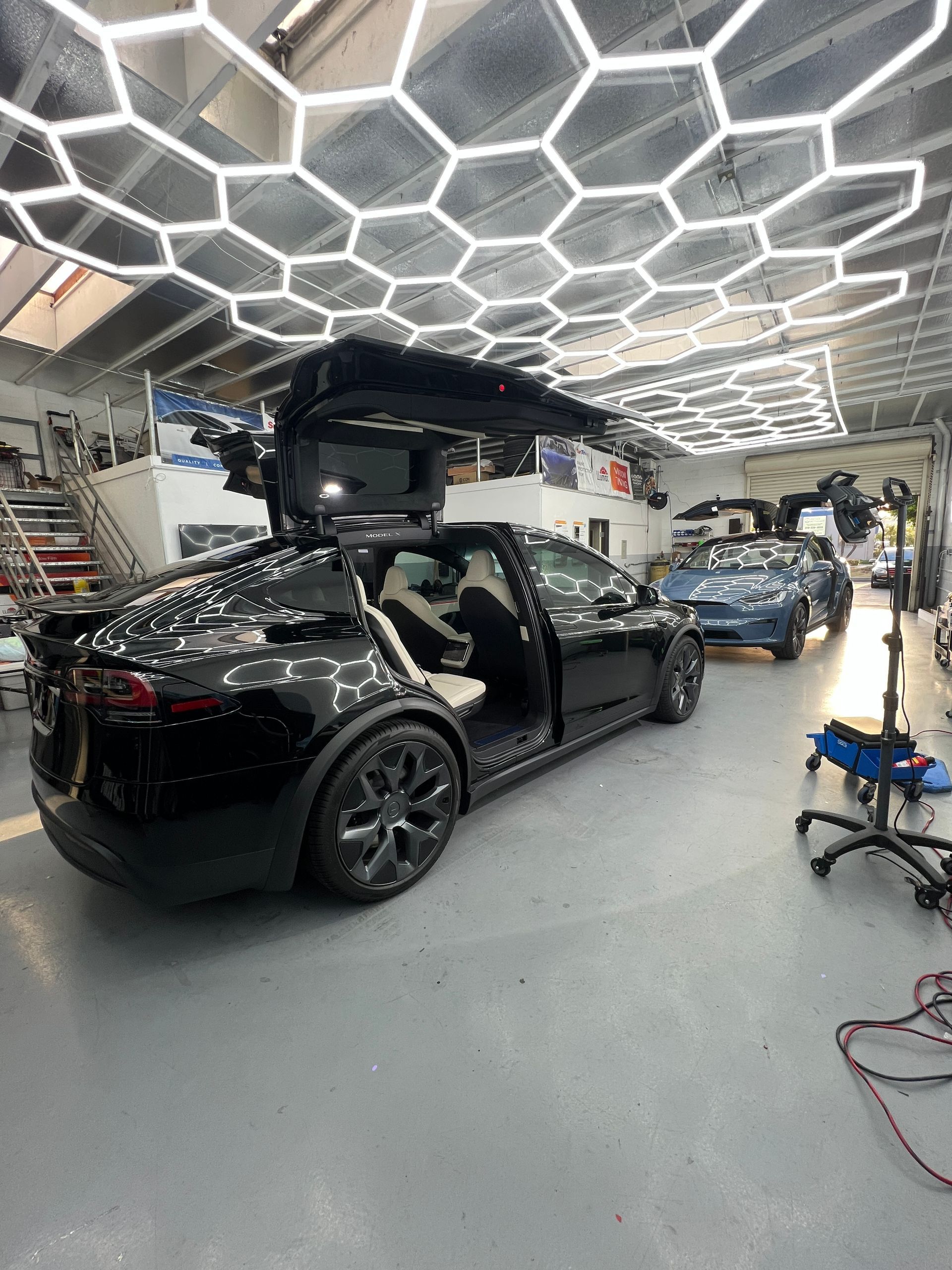 Black Tesla in garage with falcon-wing doors open under hexagonal ceiling lights