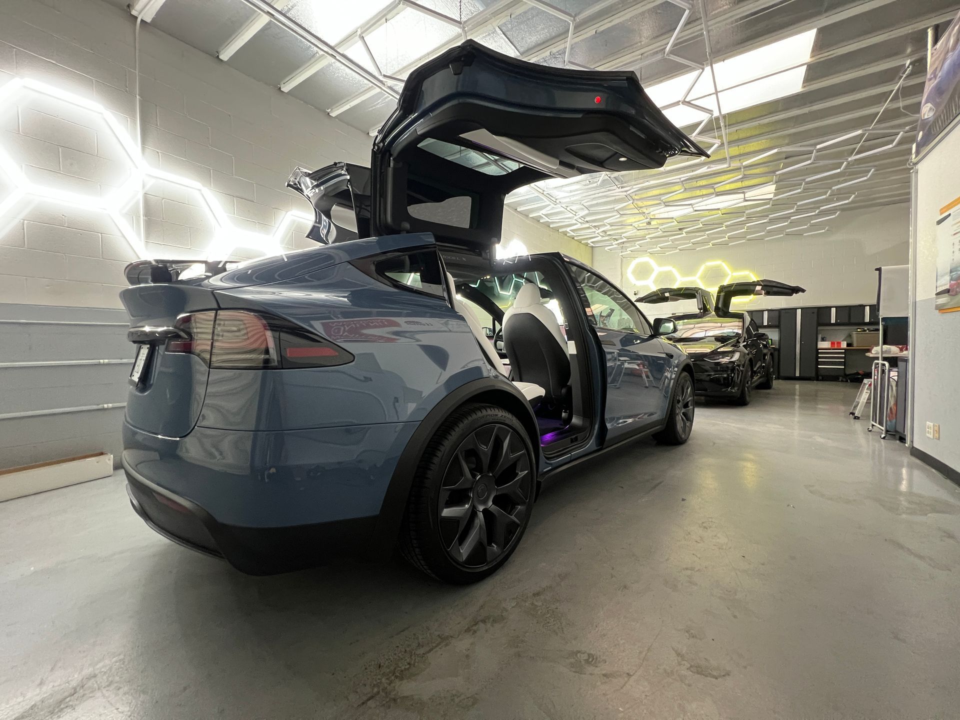 Blue SUV with gullwing doors open in a bright auto shop, with other cars in the background