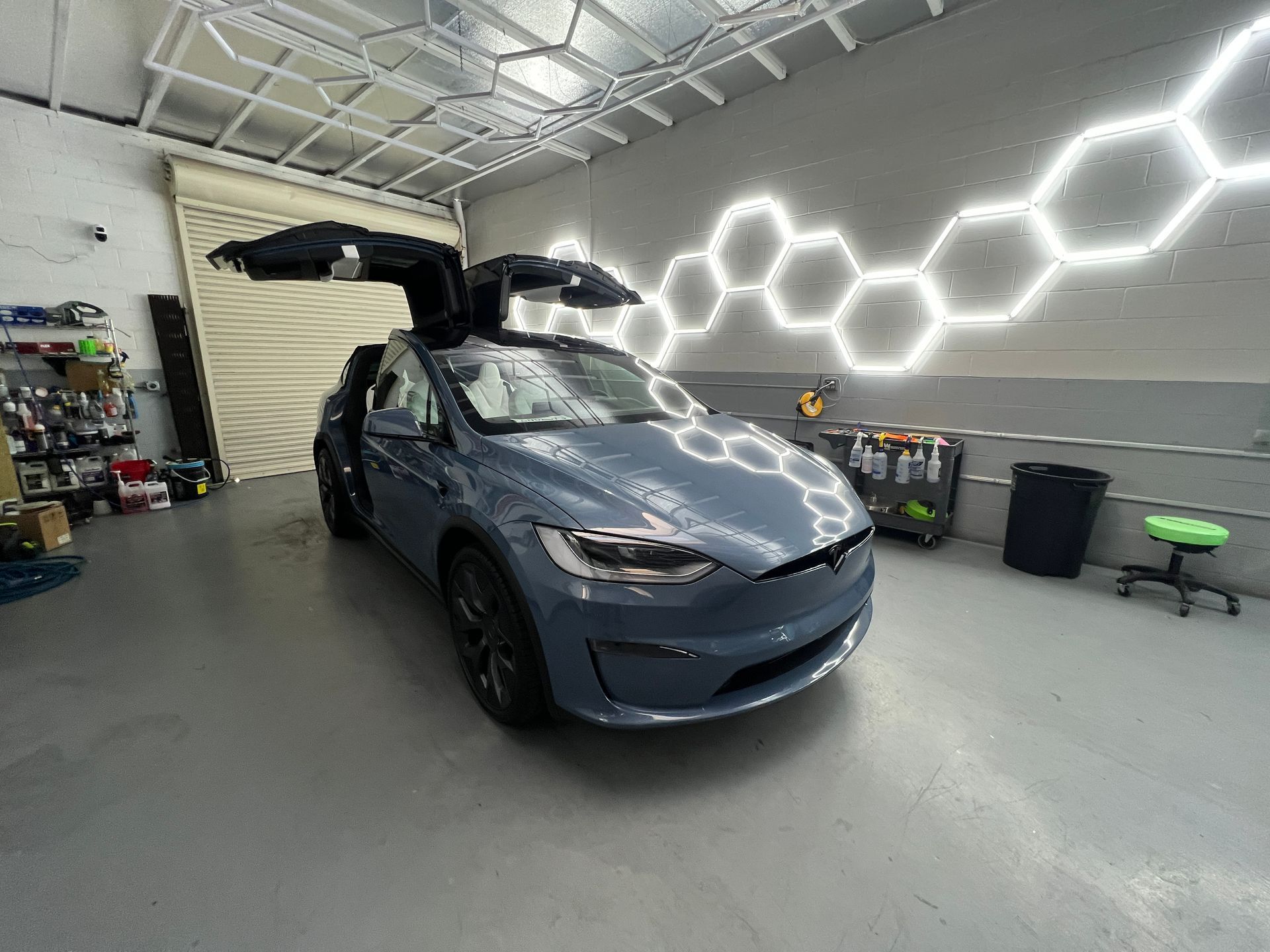 Blue Tesla with open falcon-wing door in a modern garage with hexagonal wall lights.