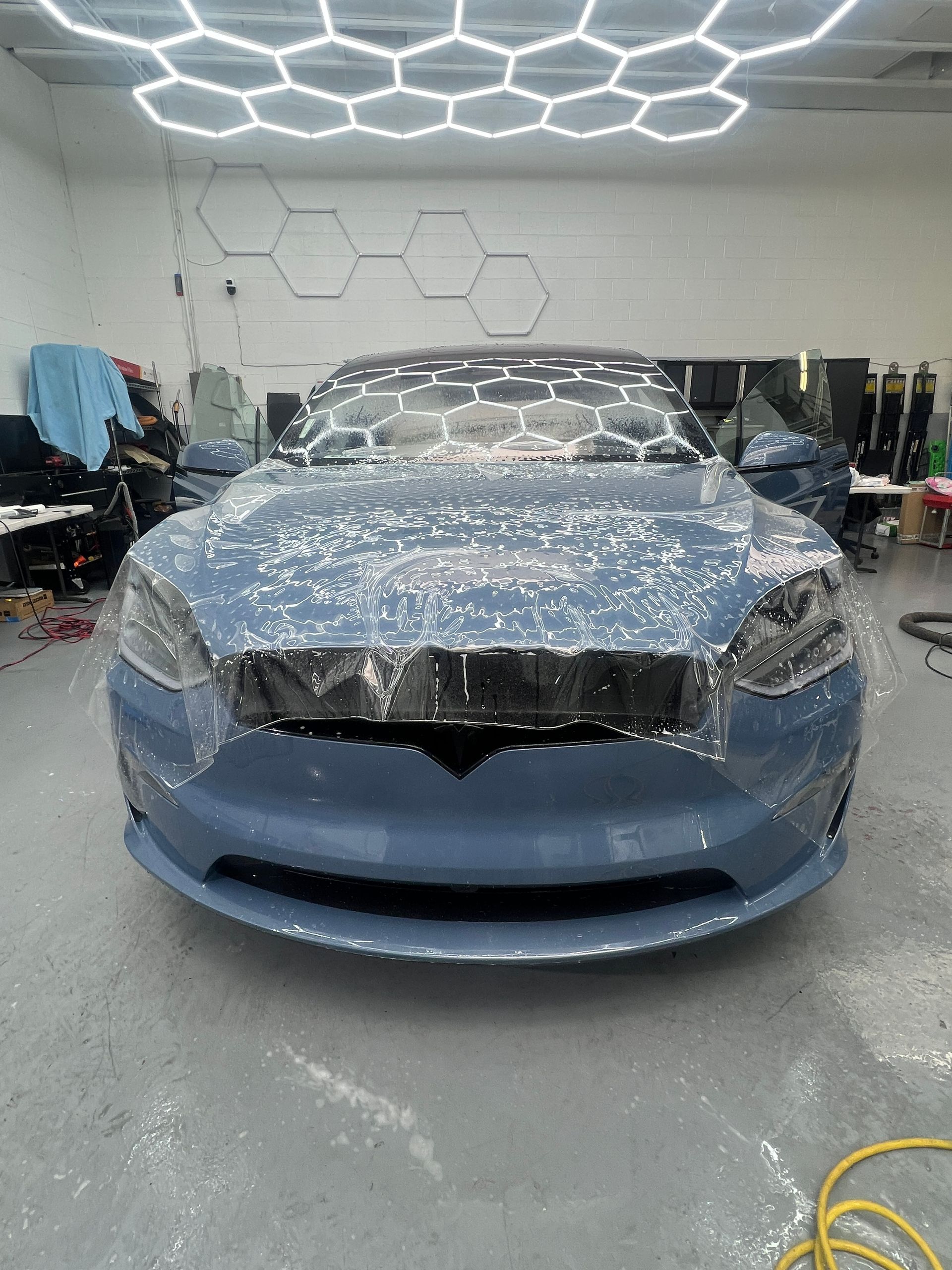Blue car covered in protective wrap inside an auto body shop, with hexagon ceiling lights overhead