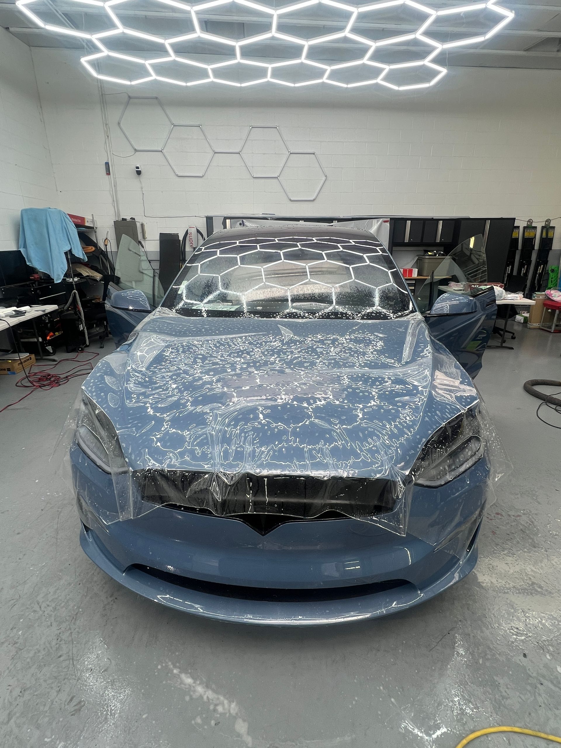 Blue car in a workshop, covered in protective wrap under bright hexagonal ceiling lights.