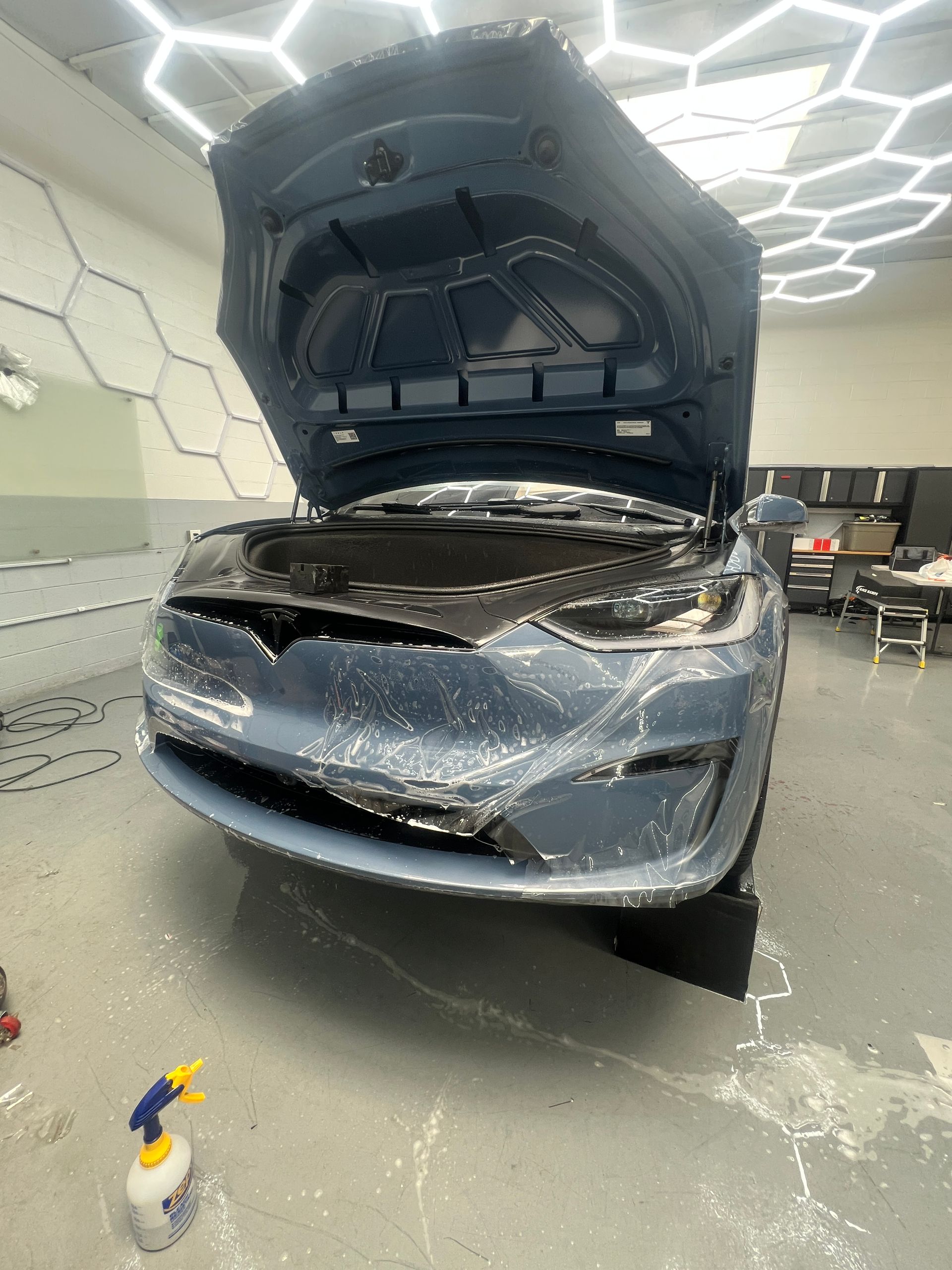 Blue car with hood open in an auto repair shop, front bumper and grille damaged.