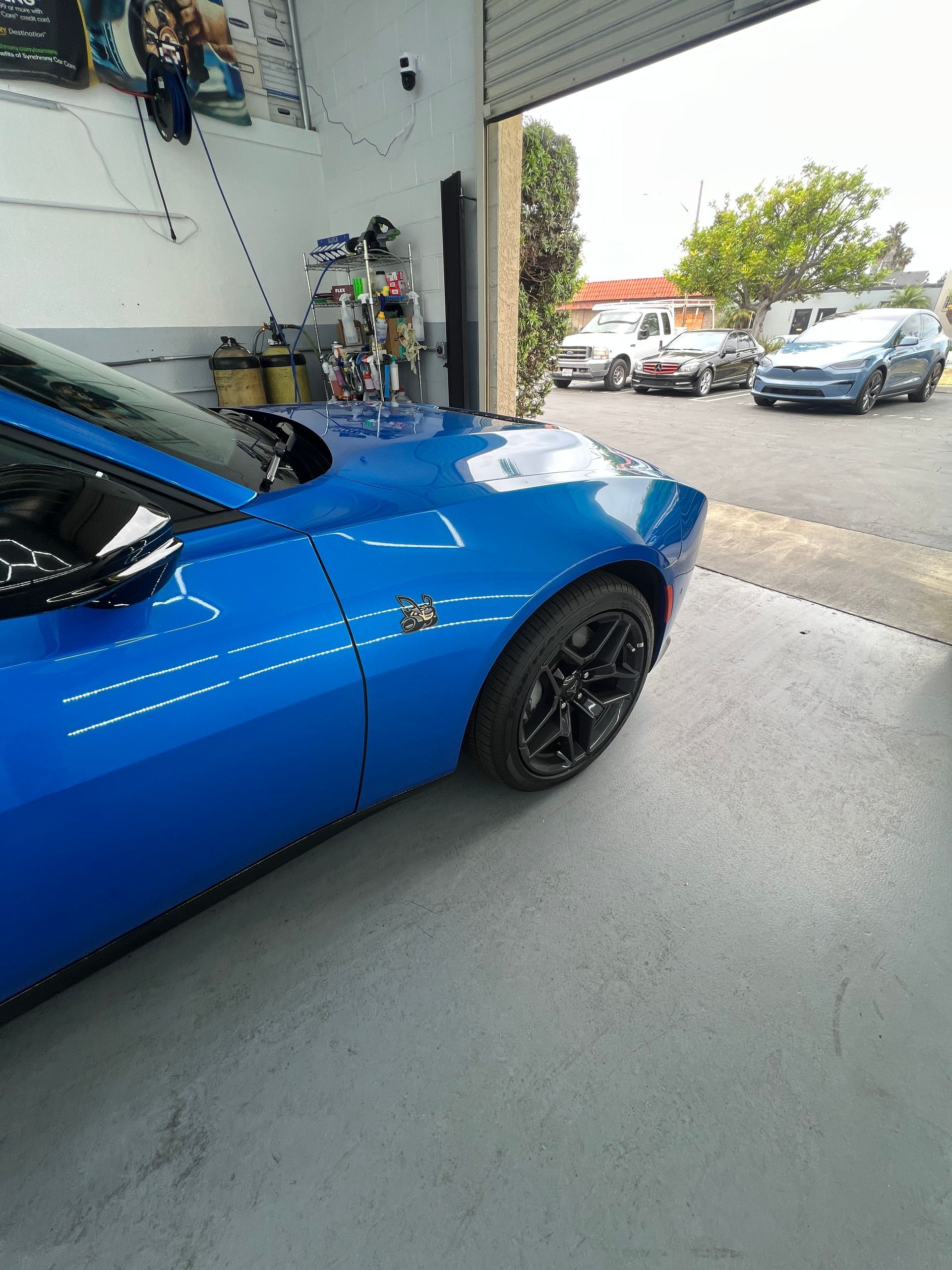 Blue sports car parked inside a garage, with a driveway and parked cars visible outside.