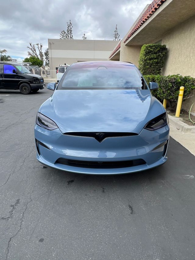 Blue Tesla parked in a lot, front view, with a beige building and hedge nearby