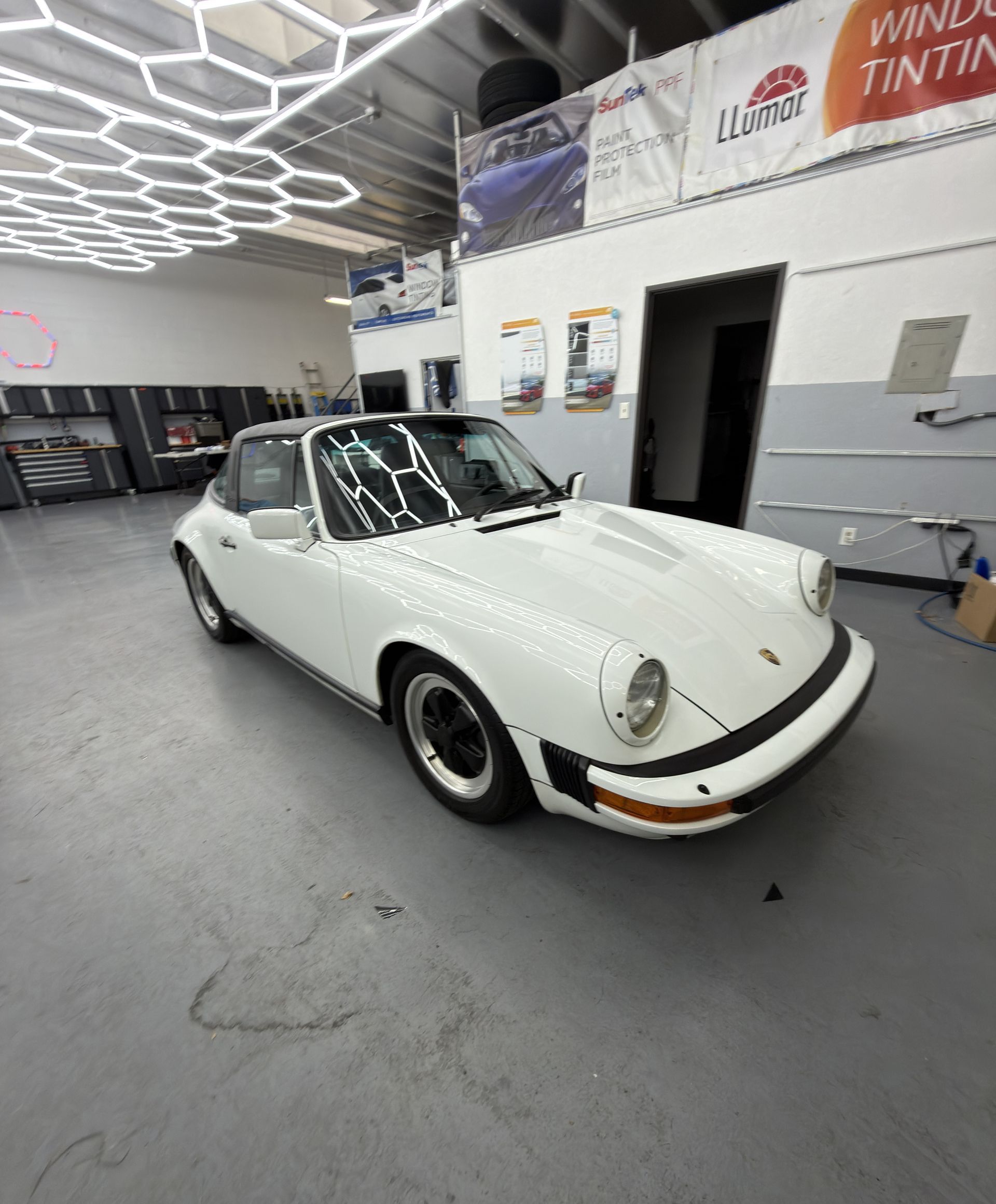 White classic Porsche 911 in an auto shop with hexagon ceiling lights and tinting signs in the background