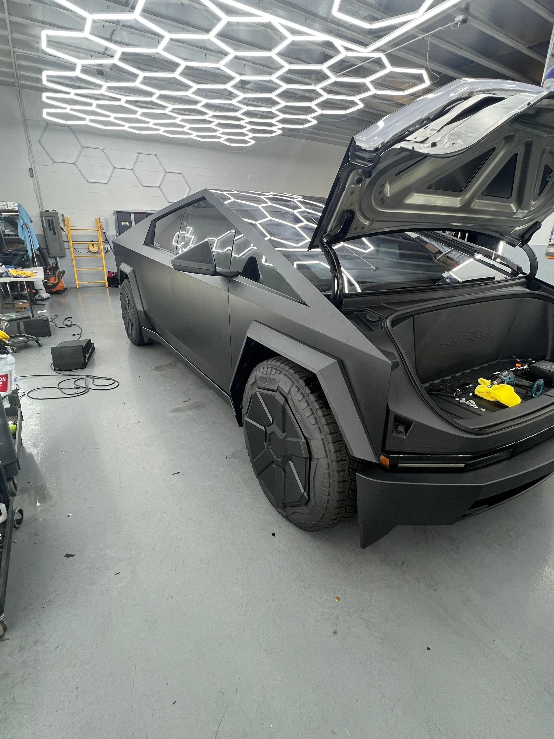 Matte black Cybertruck in a workshop, front hood open under bright hexagonal ceiling lights.