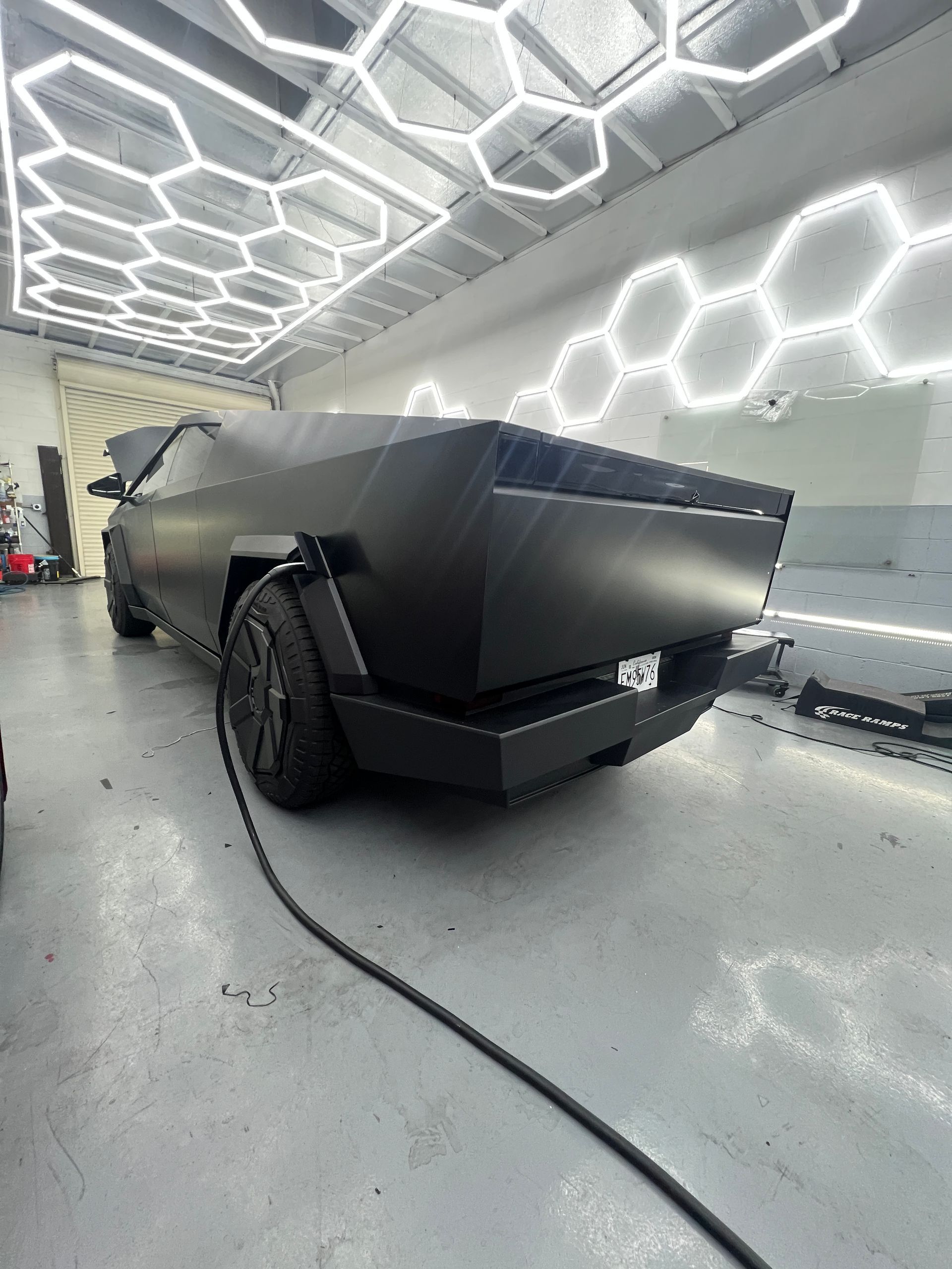 Matte black angular truck in a bright garage with hexagonal ceiling lights and a charging cable on the floor
