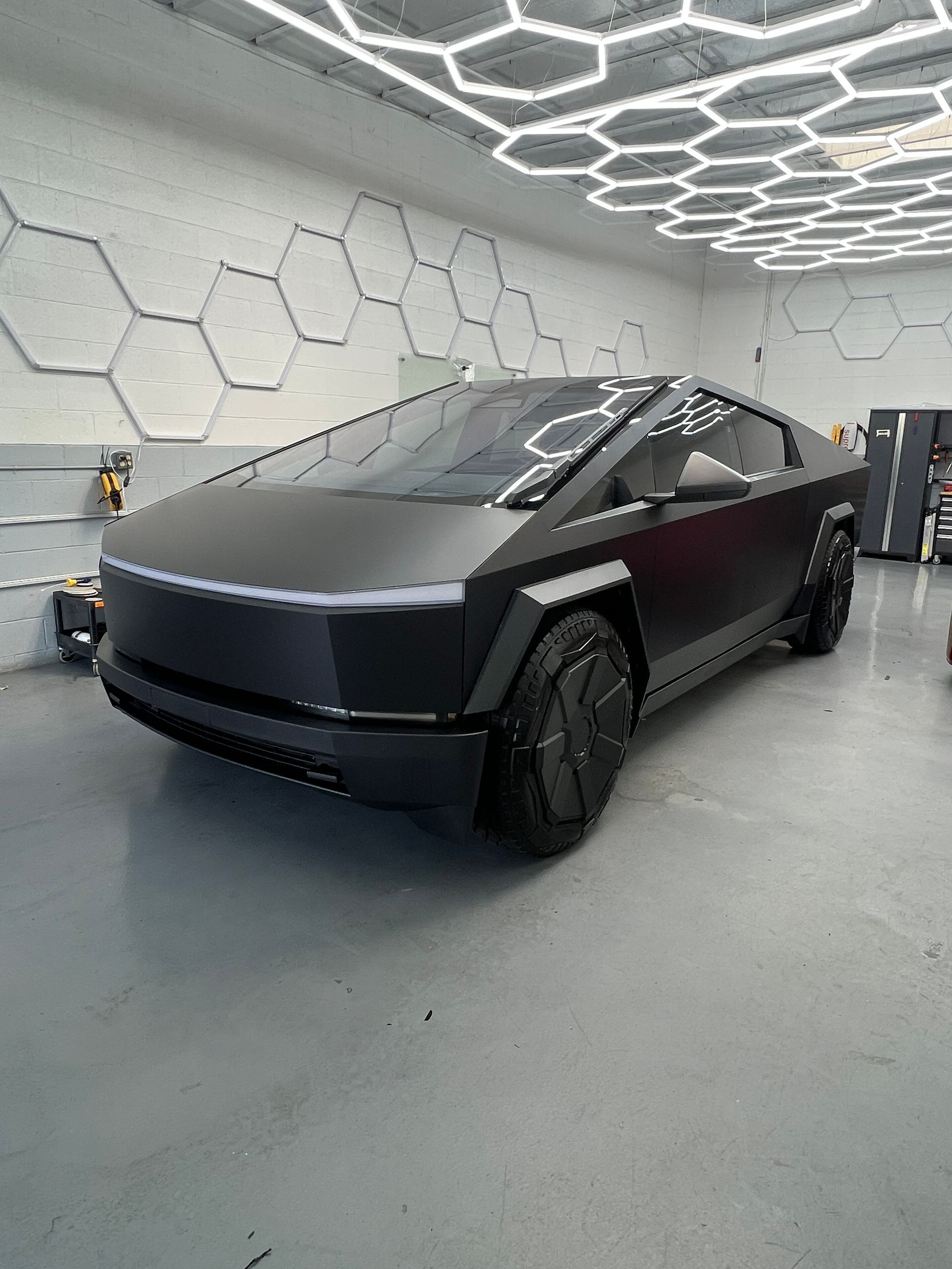 Matte black Tesla Cybertruck in a futuristic white garage with hexagonal wall panels